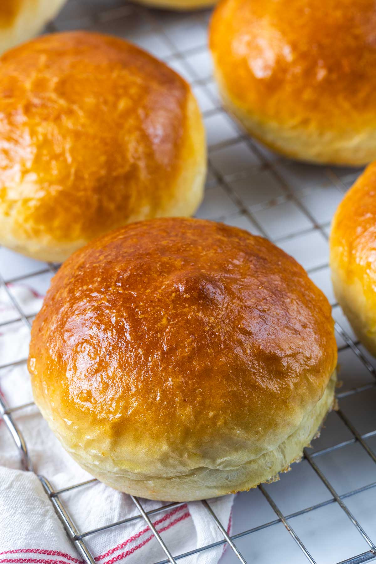 Golden brown, shiny homemade brioche buns cooling on a metal wire rack with a white and red towel underneath.