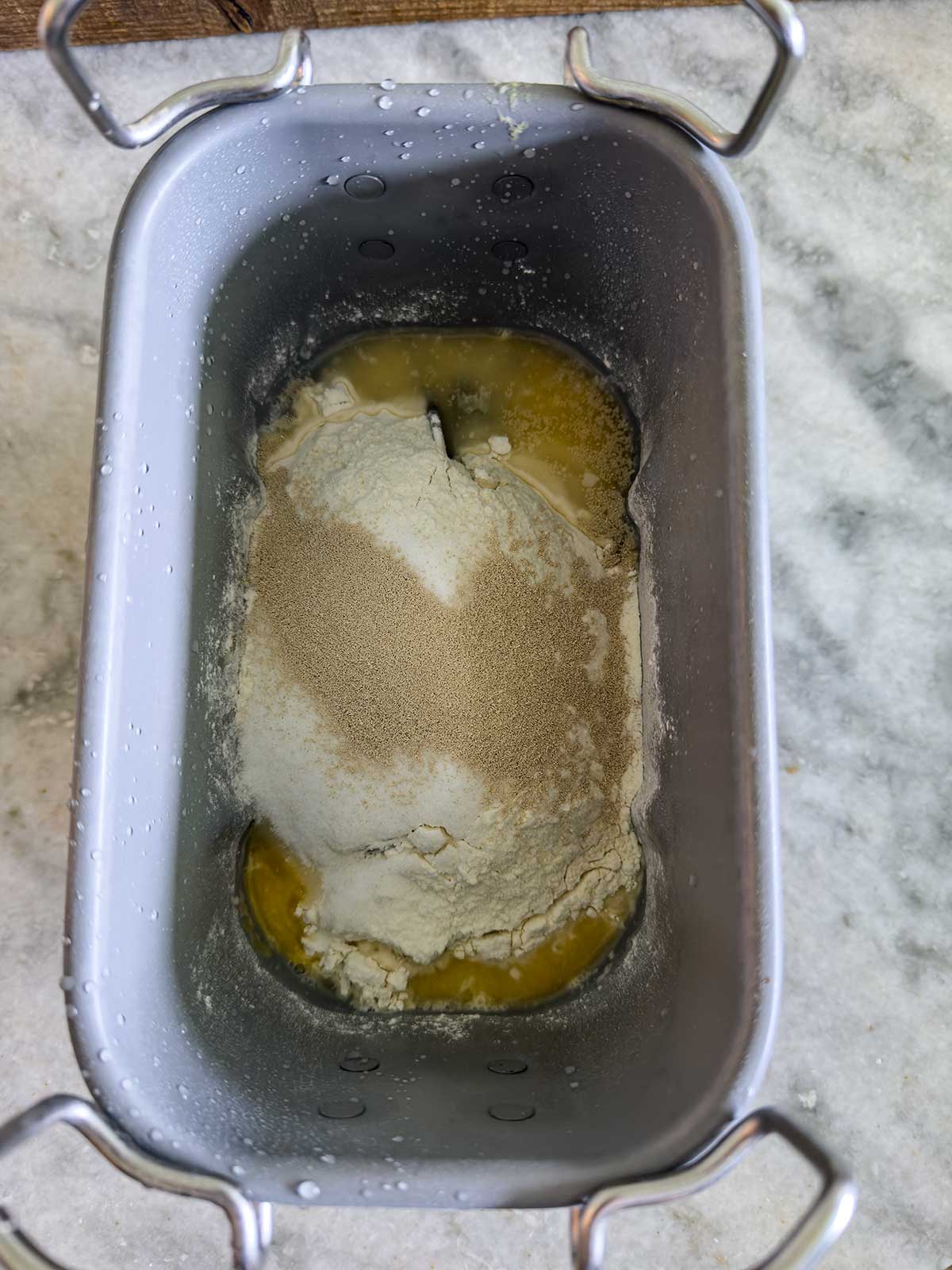 Overhead view of a bread machine pan with flour, yeast, and melted butter or oil for bread machine brioche buns, all unmixed, on a marble countertop.
