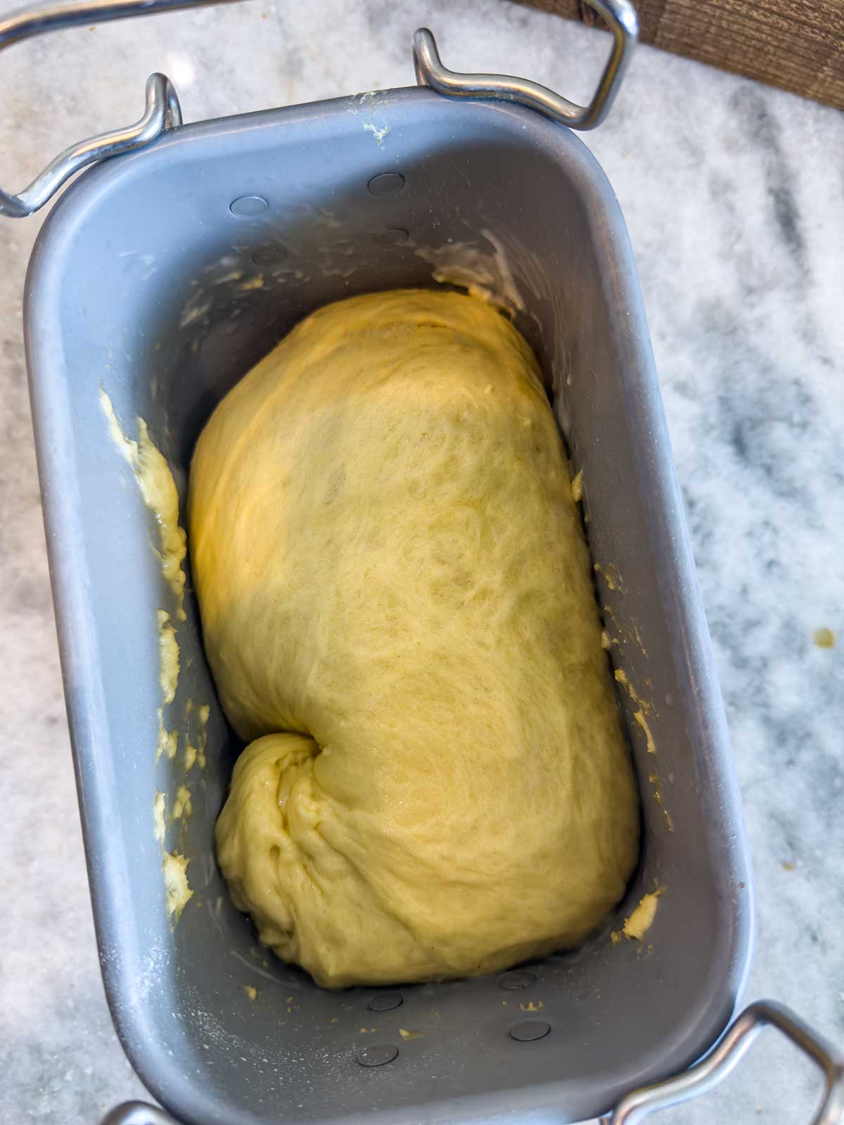 A smooth ball of yellow bread dough for bread machine brioche buns rests in a metal pan, ready for baking. The pan sits on a marble countertop.
