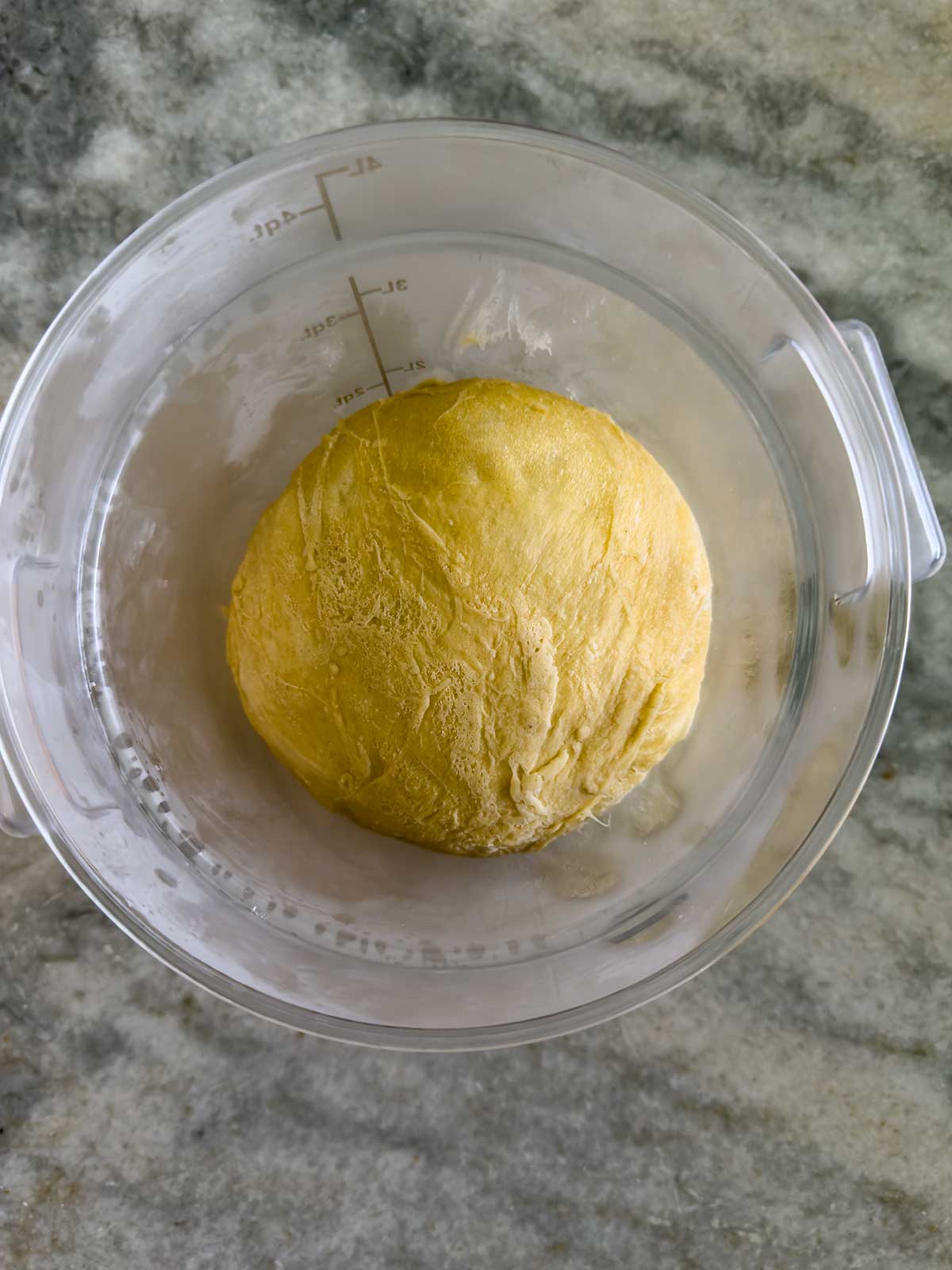 A ball of yellow dough, perfect for bread machine brioche buns, sits in a clear glass mixing bowl on a gray marble surface.