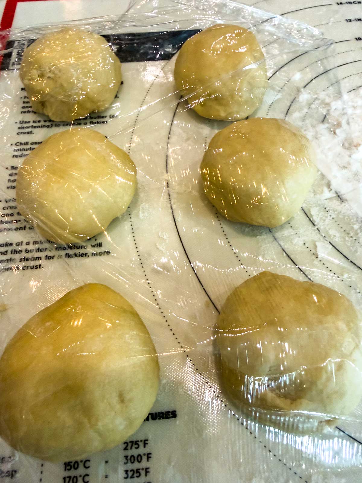 Six dough balls, likely bread machine brioche buns, rest under plastic wrap on a floured surface, proofing before baking. Temperature and baking instructions are partially visible on the mat underneath.
