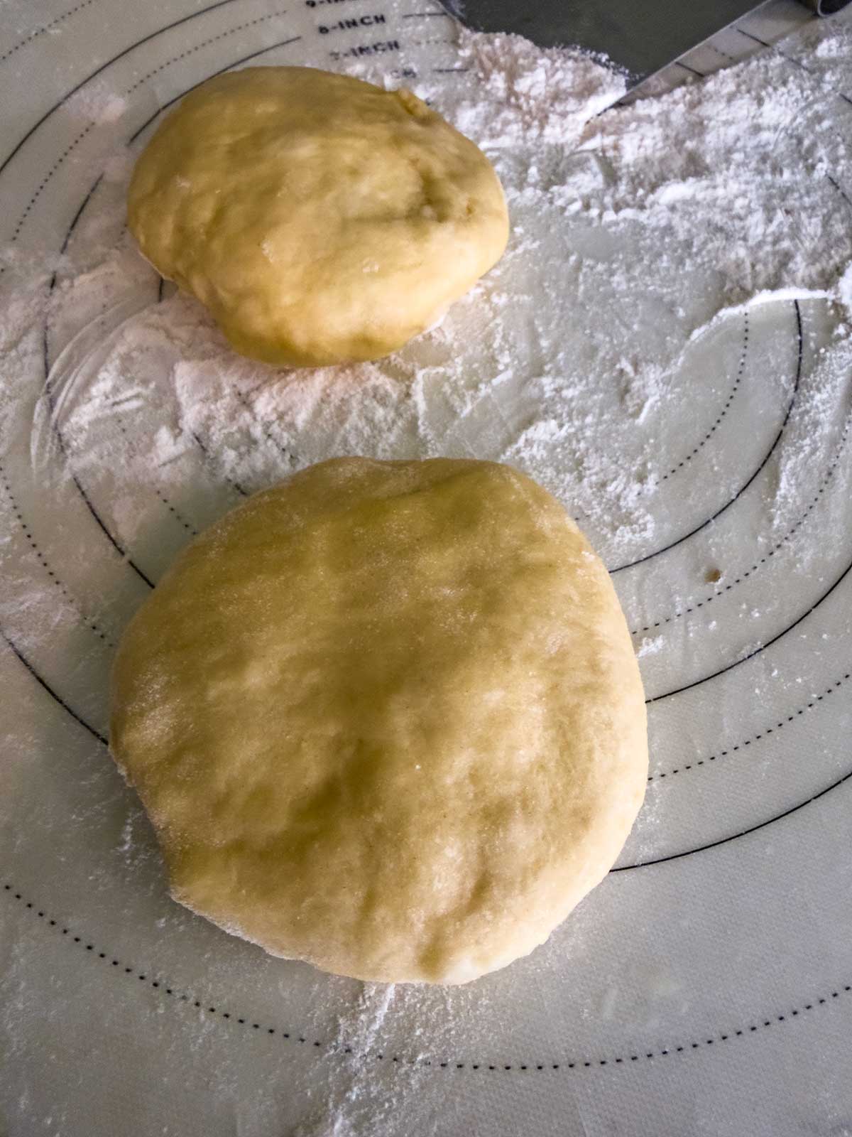 Two balls of dough for bread machine brioche buns rest on a floured pastry mat with measurement markings, ready for baking or further preparation. A dough cutter is partially visible in the upper right corner.