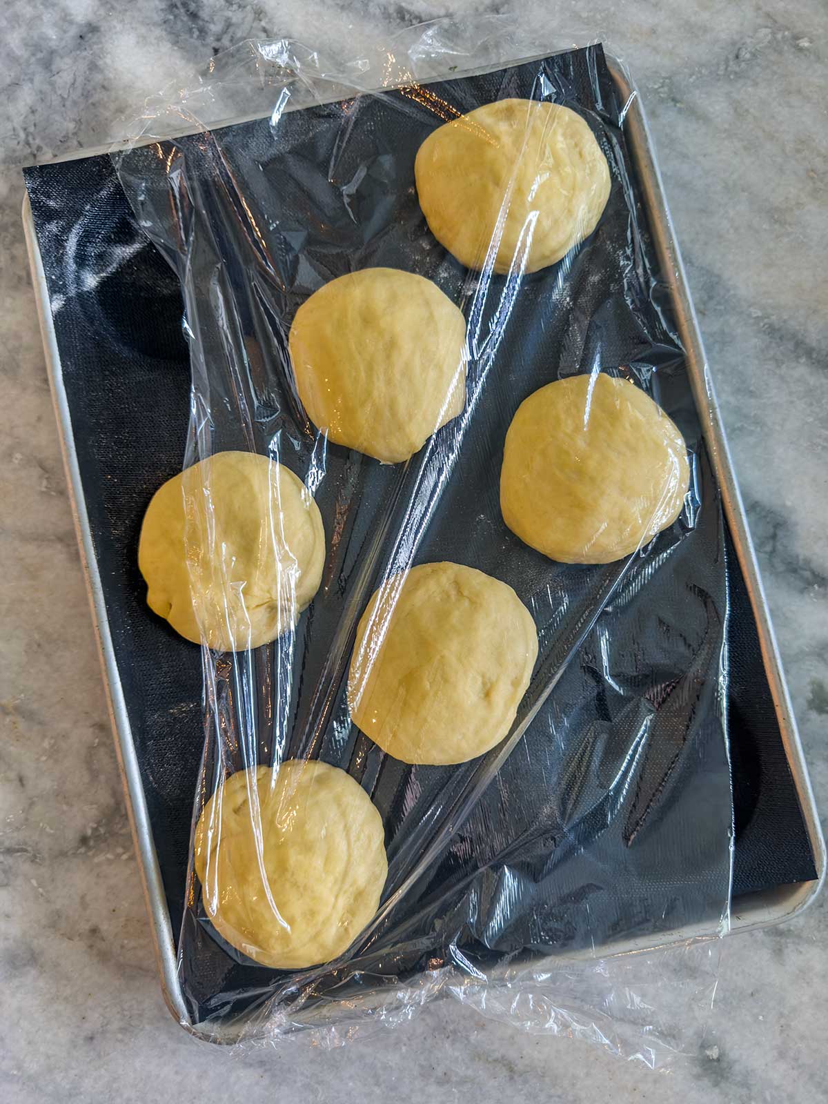 Five round bread machine brioche buns rest on a black baking tray, covered loosely with plastic wrap, atop a marble countertop.