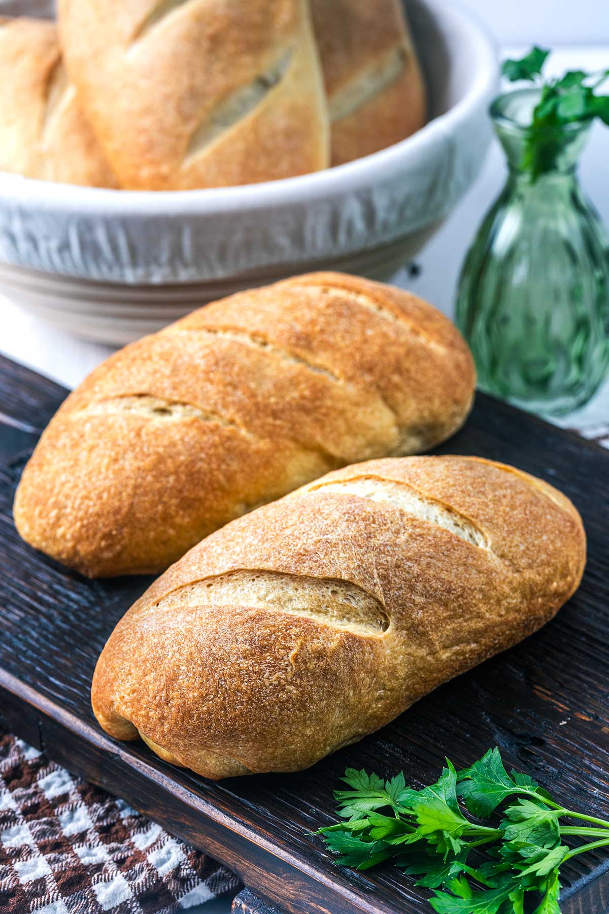 Two golden brown baguettes with slash marks rest on a dark wooden board, with fresh parsley nearby. A white bowl with more bread and a green glass bottle are in the background. A plaid cloth is partially visible.