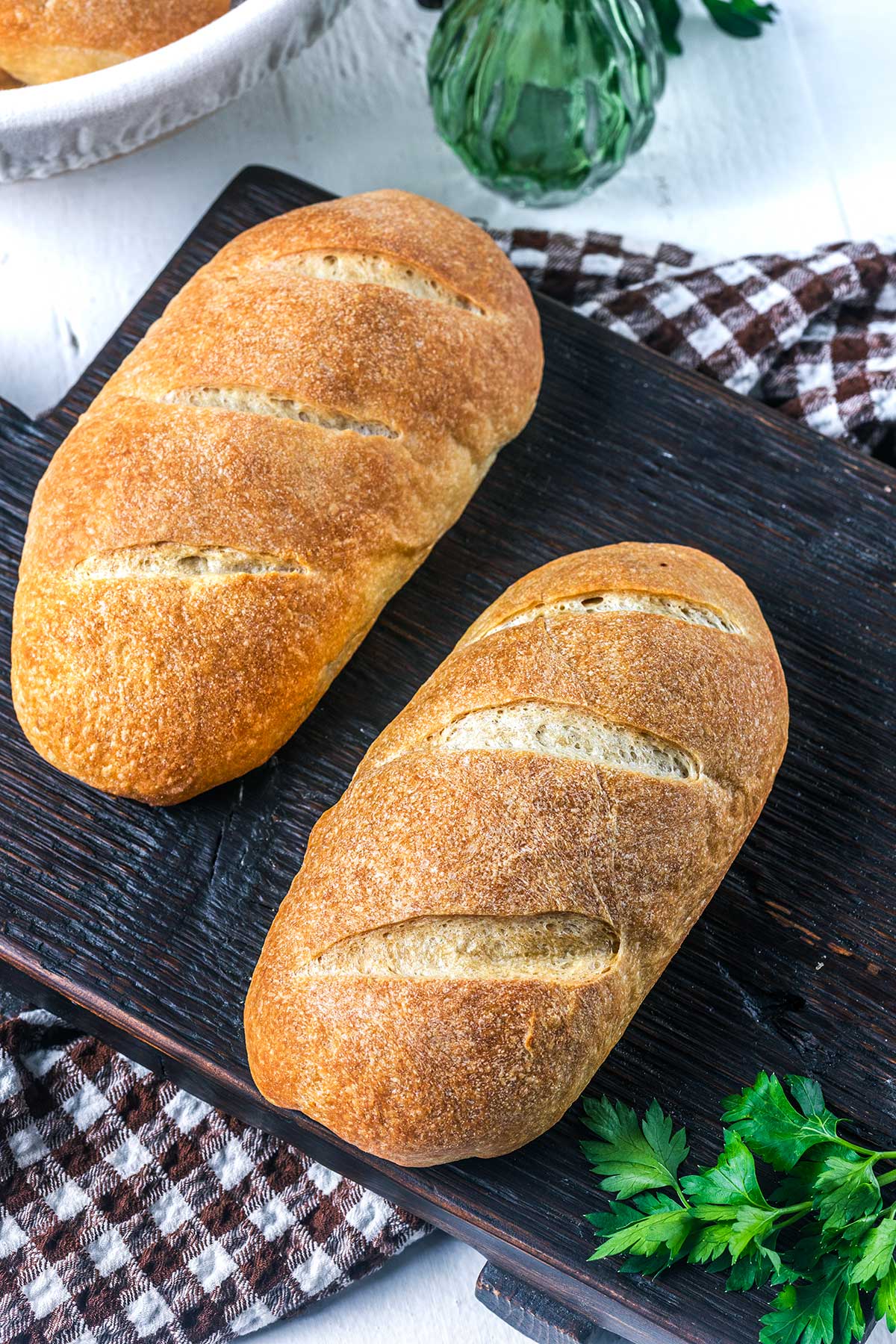 Two golden-brown loaves of bread with slashed tops rest on a dark wooden board. A checkered cloth and fresh parsley are nearby, with part of a green glass bottle visible in the background.