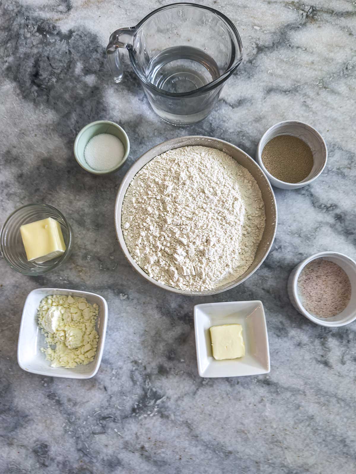 A marble countertop displays bowls containing flour, water, yeast, sugar, butter, powdered milk, and salt, all arranged neatly and ready for baking preparation.