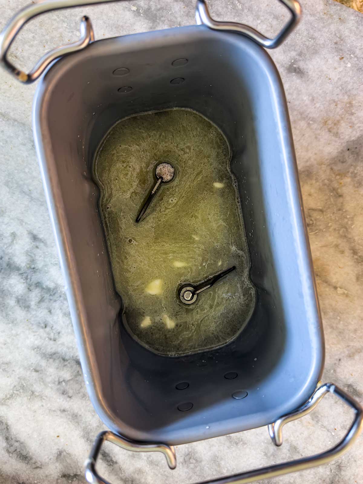 A bread machine pan with two kneading paddles, containing melted butter and minced garlic on the bottom, placed on a marble countertop.