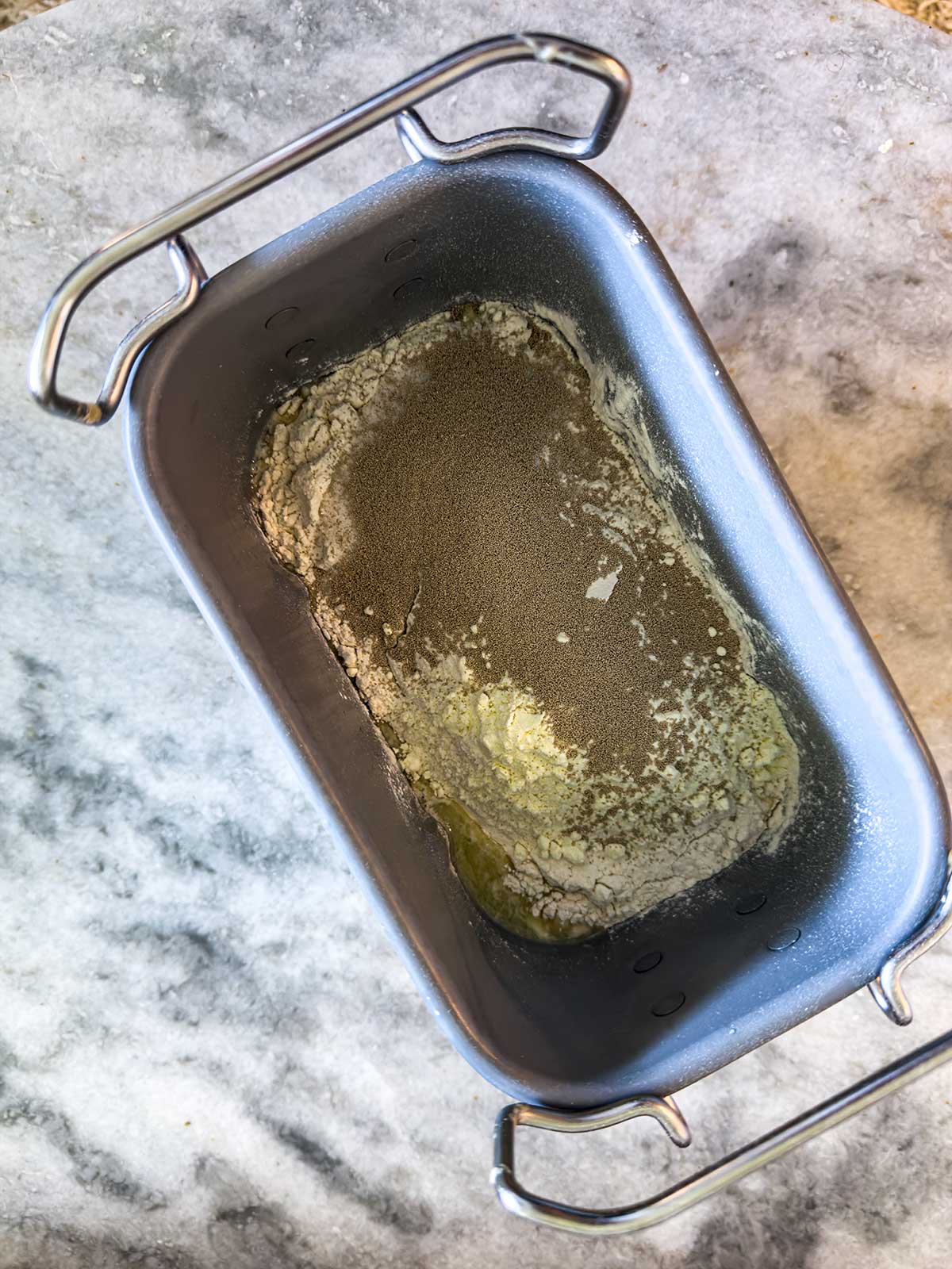 A bread machine pan with flour, yeast, and melted butter on a marble countertop, ready for mixing.