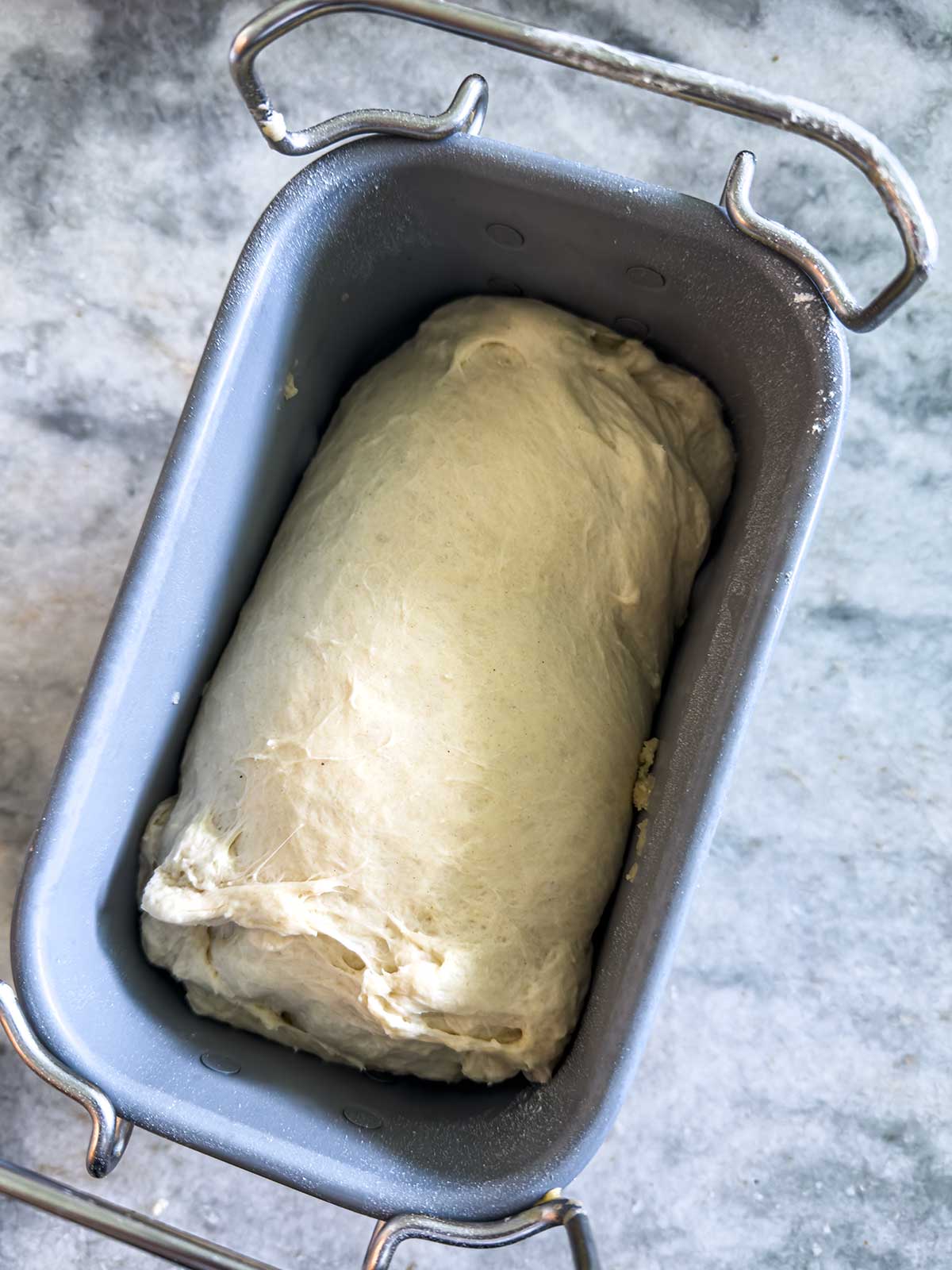 Unbaked bread dough shaped into a loaf sits in a metal bread pan on a gray, textured surface.