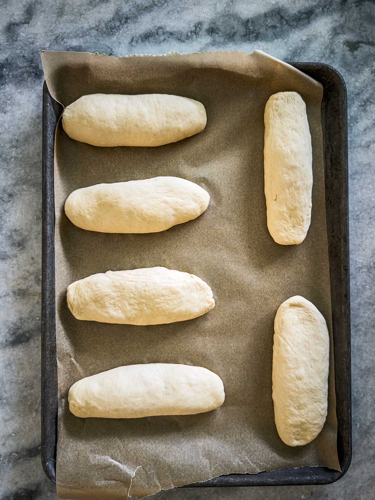 Six unbaked bread rolls are evenly spaced on a parchment-lined baking tray, placed on a gray marble surface. The dough is smooth and oval-shaped, ready for baking.