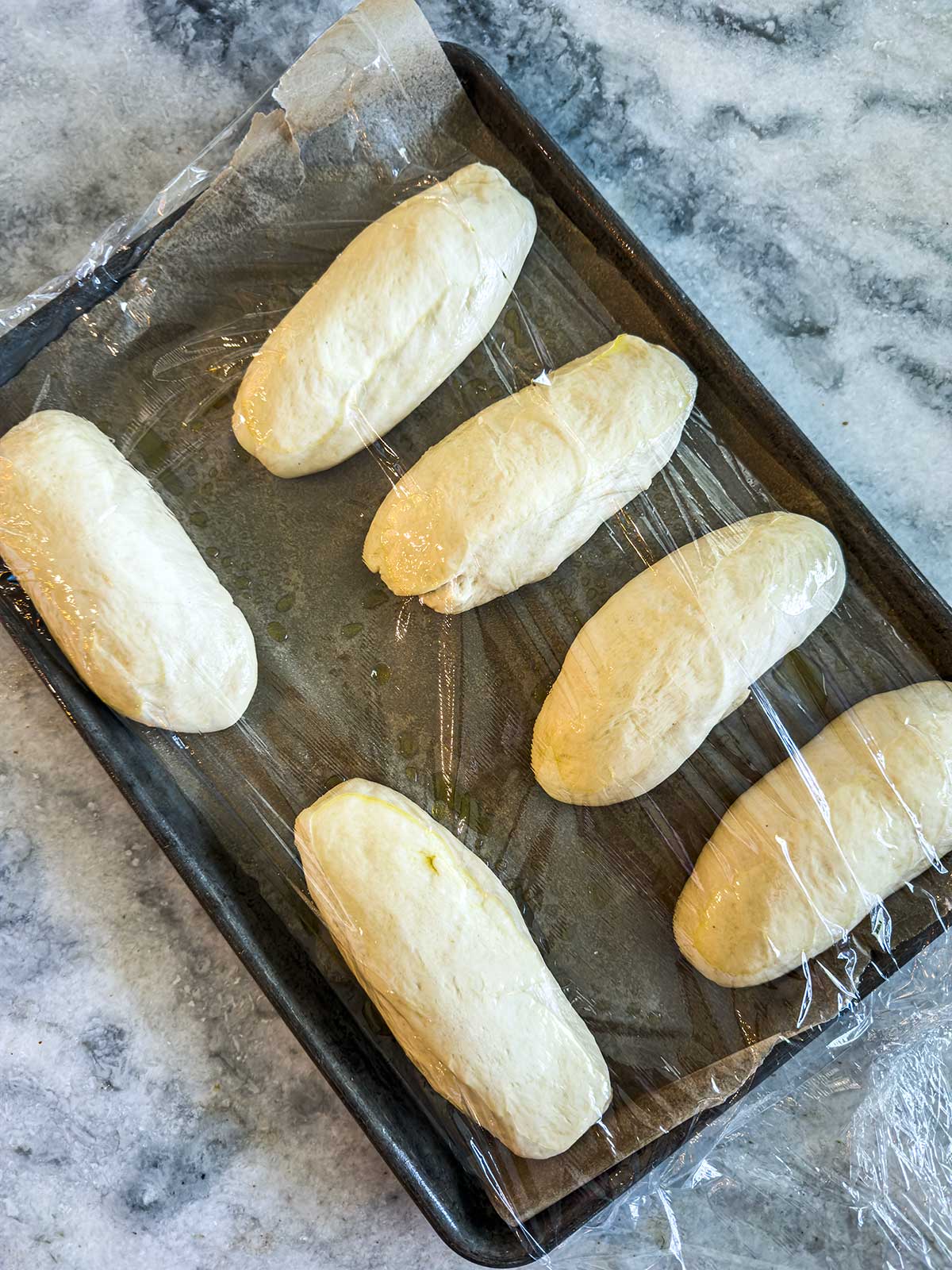 Six unbaked bread rolls are placed on a baking sheet, covered loosely with plastic wrap, on a marble countertop. The dough is rising before baking.