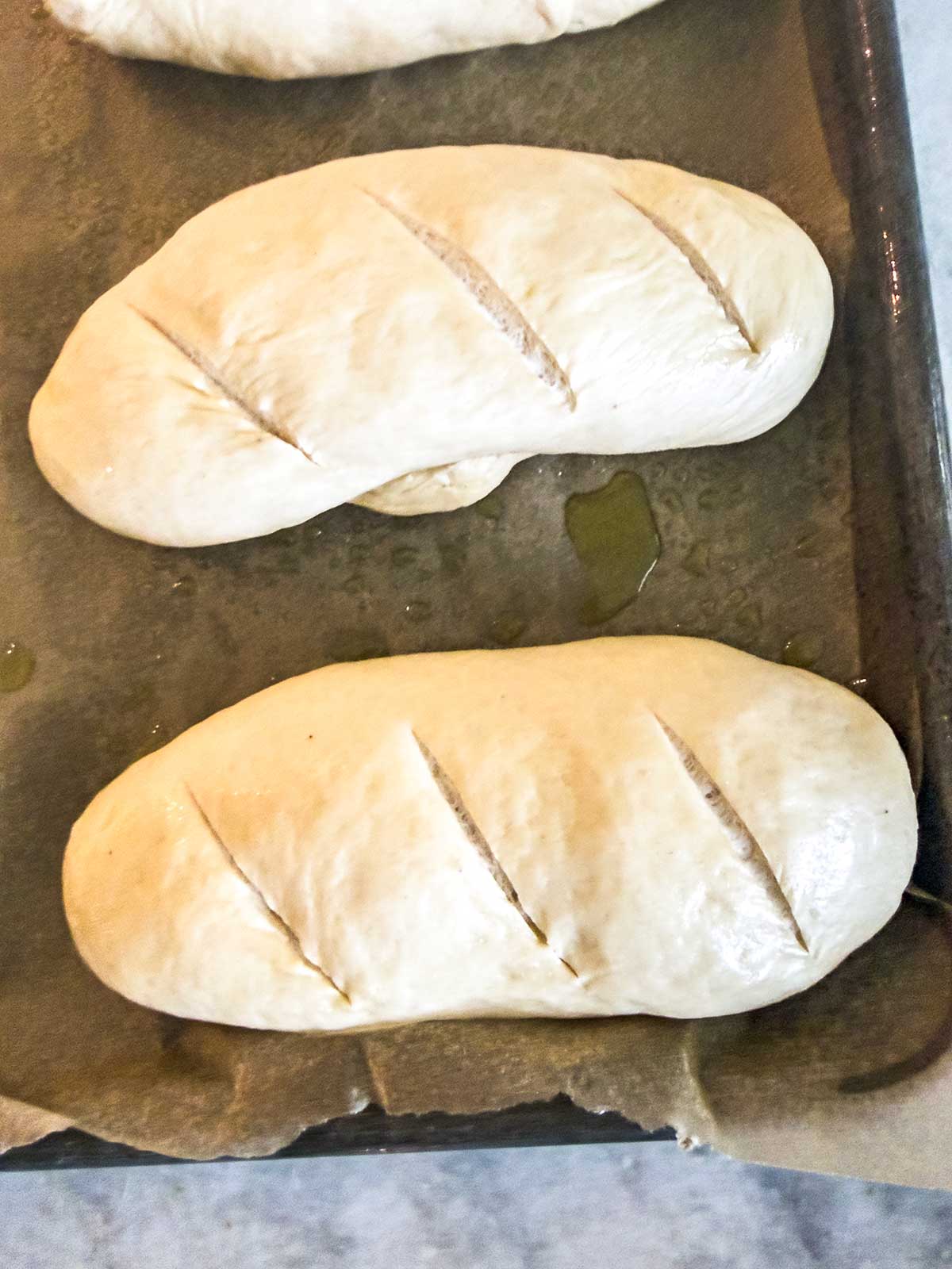 Two unbaked loaves of bread dough with three diagonal slashes each, resting on parchment paper on a baking tray, ready to be baked.
