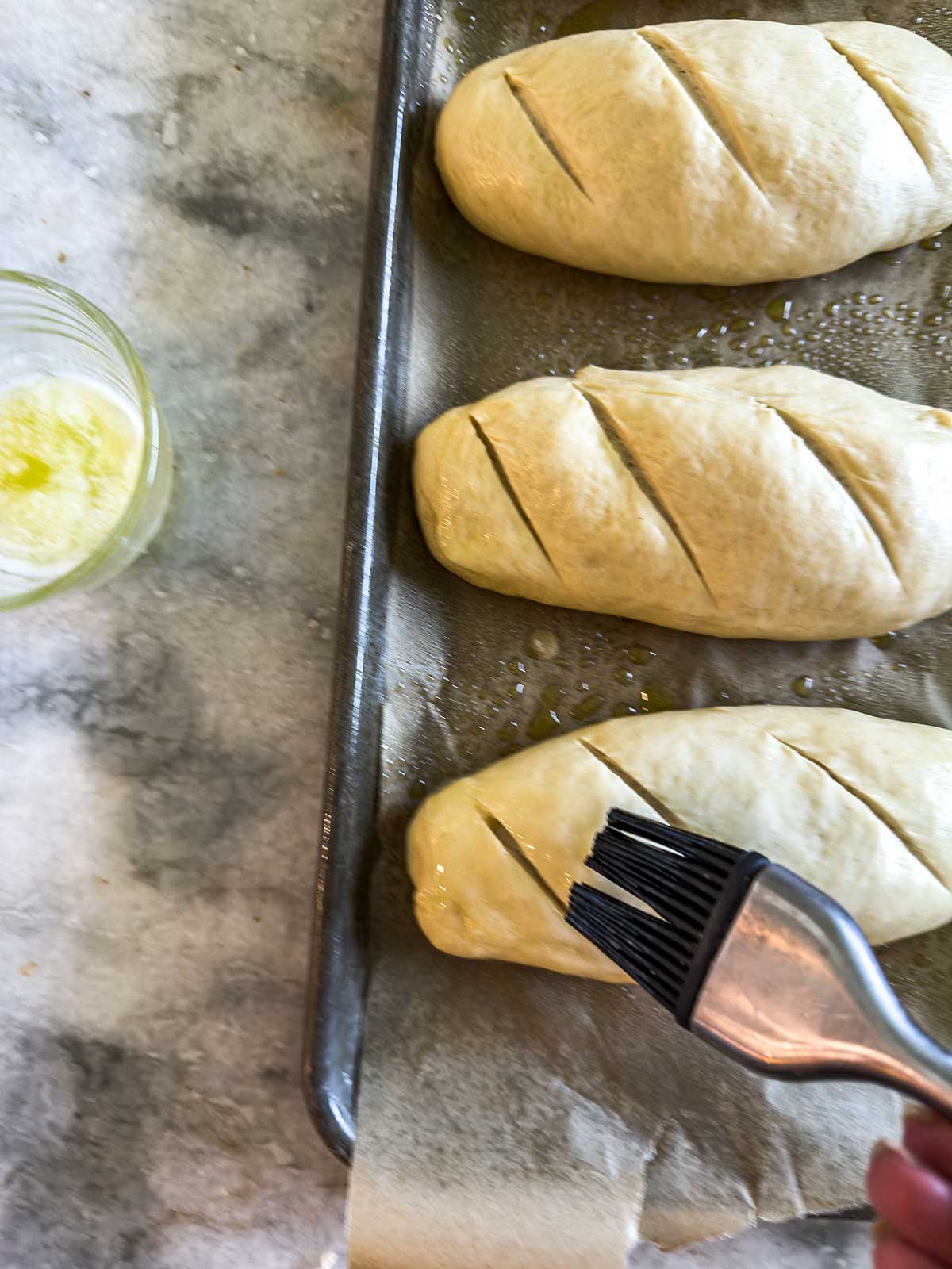 A hand uses a pastry brush to apply butter to three unbaked loaves of bread dough with diagonal slashes, placed on a parchment-lined baking sheet. A glass of melted butter sits nearby on a marble counter.