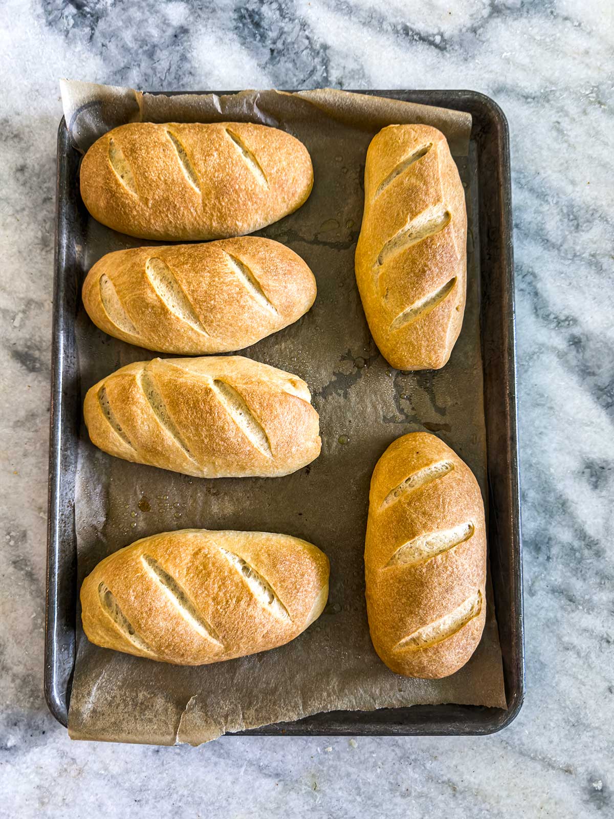 Six golden brown, homemade bread rolls with diagonal slashes on top are arranged on a parchment-lined baking sheet, resting on a marble countertop.