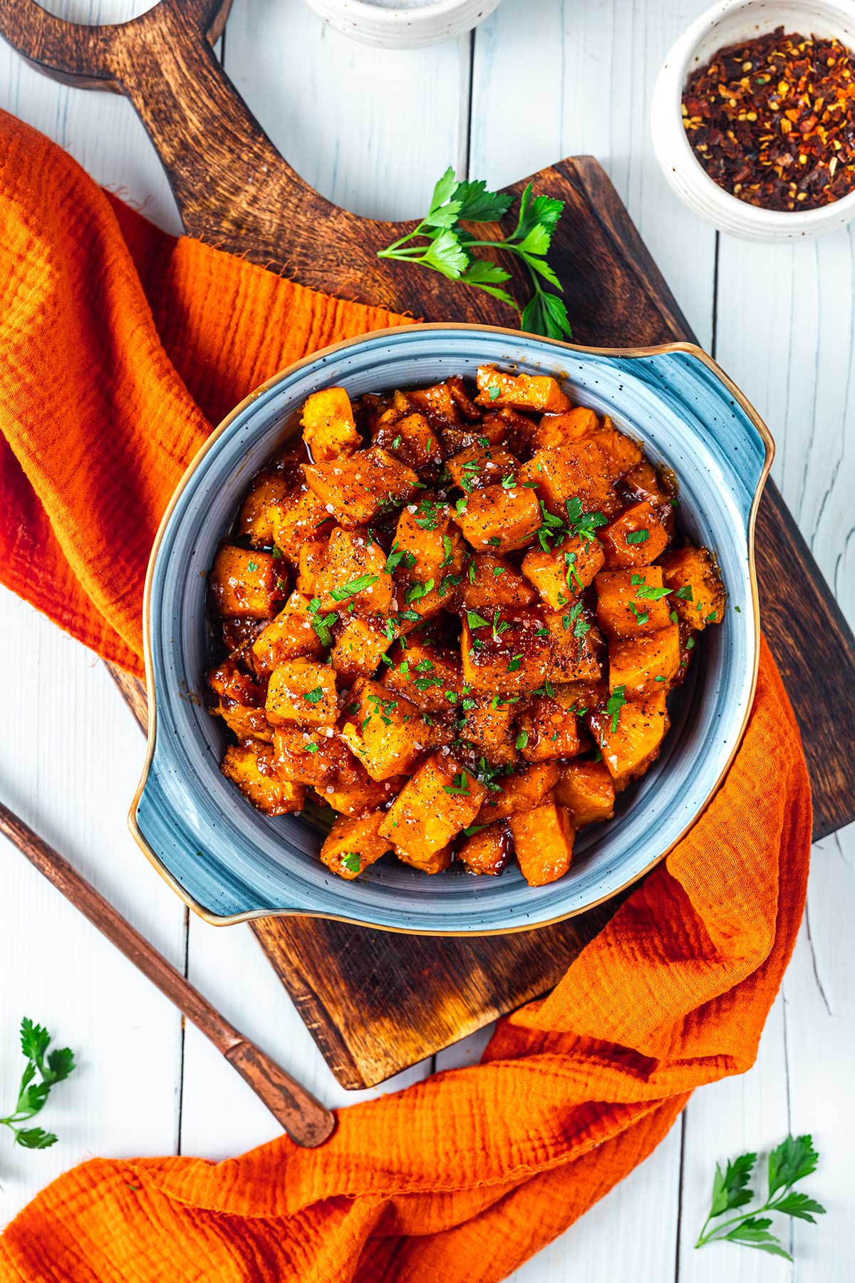 A bowl of seasoned, roasted diced potatoes garnished with chopped parsley, placed on a wooden board with an orange cloth and a small bowl of red pepper flakes nearby.