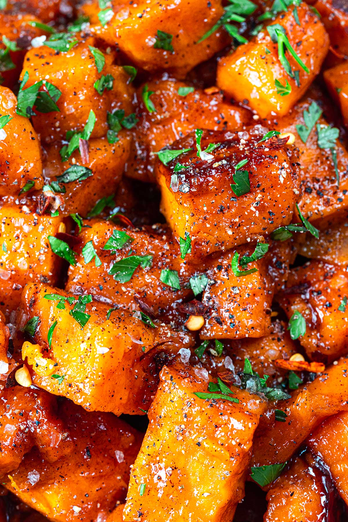 Close-up of roasted sweet potato cubes seasoned with black pepper, herbs, and coarse salt, garnished with chopped fresh parsley. The potatoes are caramelized and richly colored.