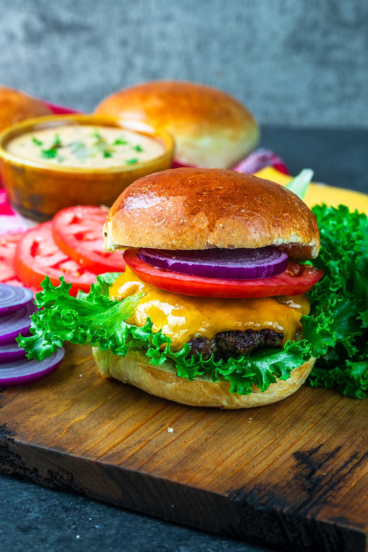 A cheeseburger with lettuce, tomato, red onion, and cheddar cheese on a wooden board, with sliced tomatoes, onions, sauce, and extra buns in the background.