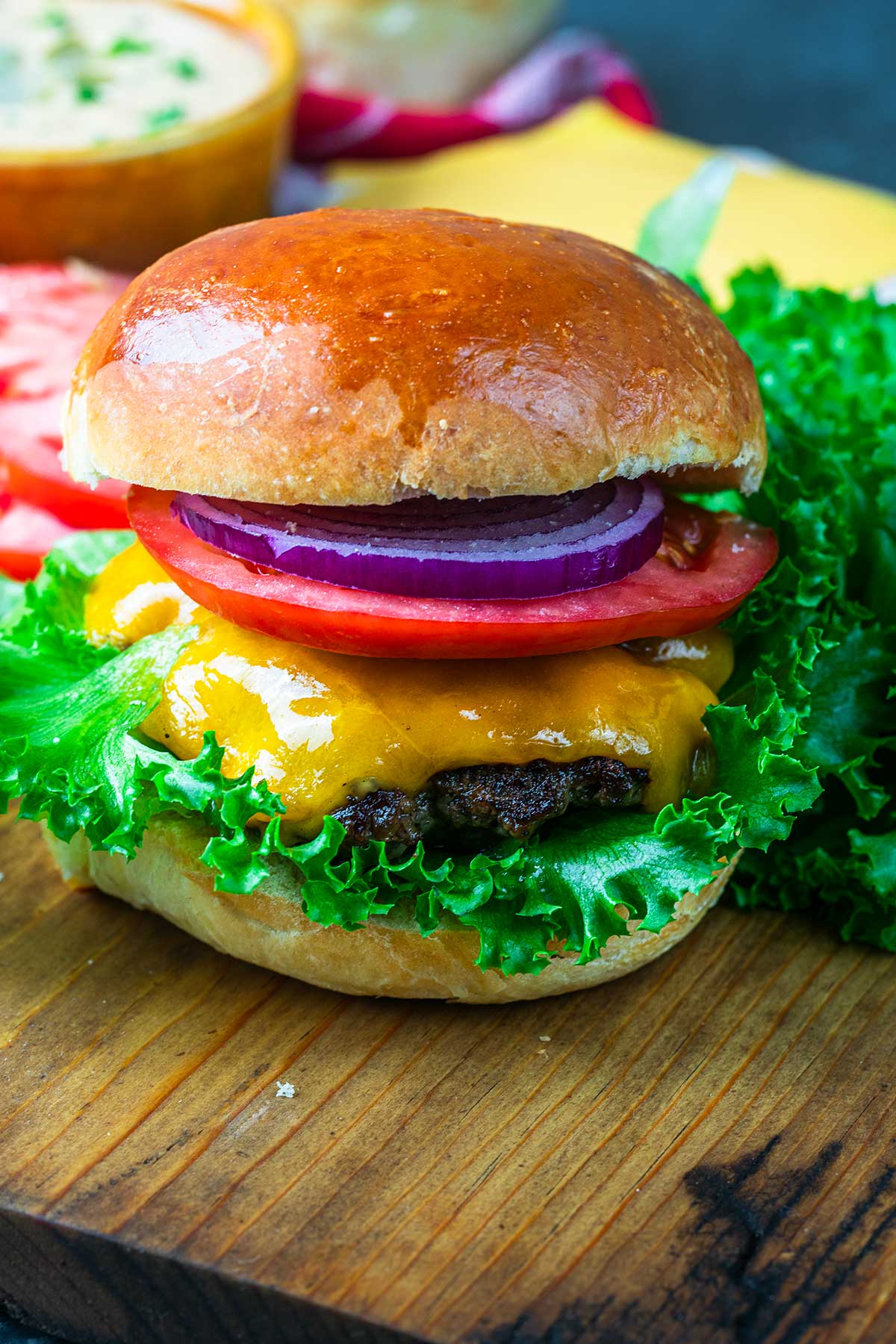 A cheeseburger on a wooden board, topped with leafy lettuce, melted cheddar cheese, a slice of tomato, and red onion, served in a shiny, golden-brown bun.