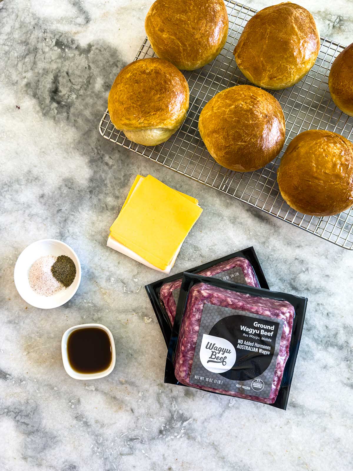 Burger buns on a cooling rack, slices of cheddar cheese, ground Wagyu beef packages, a small bowl of seasoning, and a bowl of sauce are arranged on a marble surface—perfect ingredients for mouthwatering cast iron burgers.