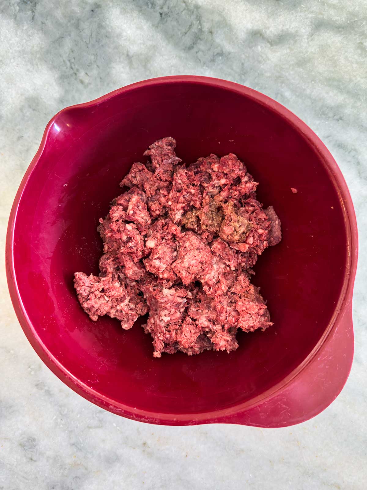 Ground beef mixture in a large red mixing bowl, sitting on a gray marble countertop, ready to be shaped into cast iron burgers.