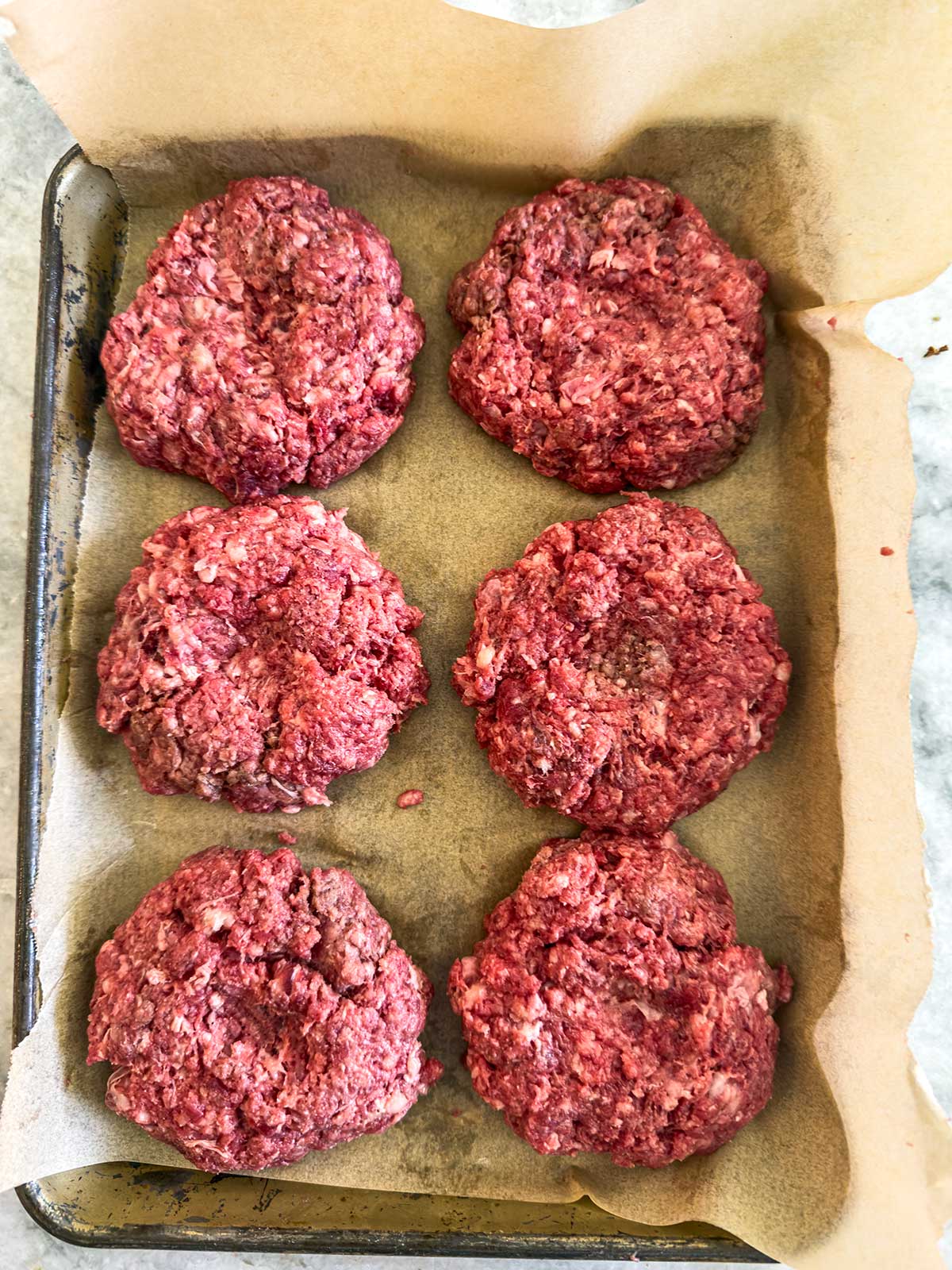Six raw beef burger patties are arranged on a parchment-lined baking tray, ready to become delicious cast iron burgers. The patties are unevenly shaped and have a coarse, textured appearance.