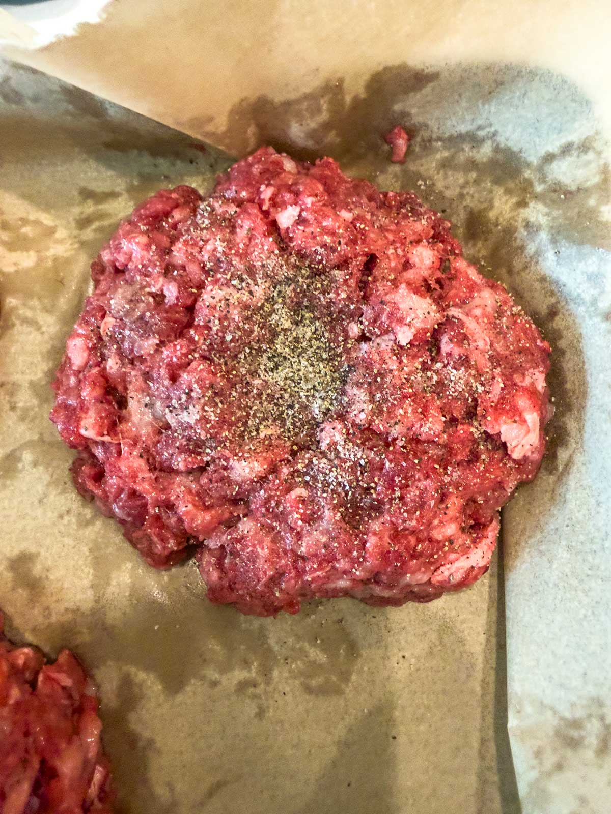 A raw hamburger patty seasoned with black pepper sits on brown parchment paper, ready to be cooked into juicy cast iron burgers.