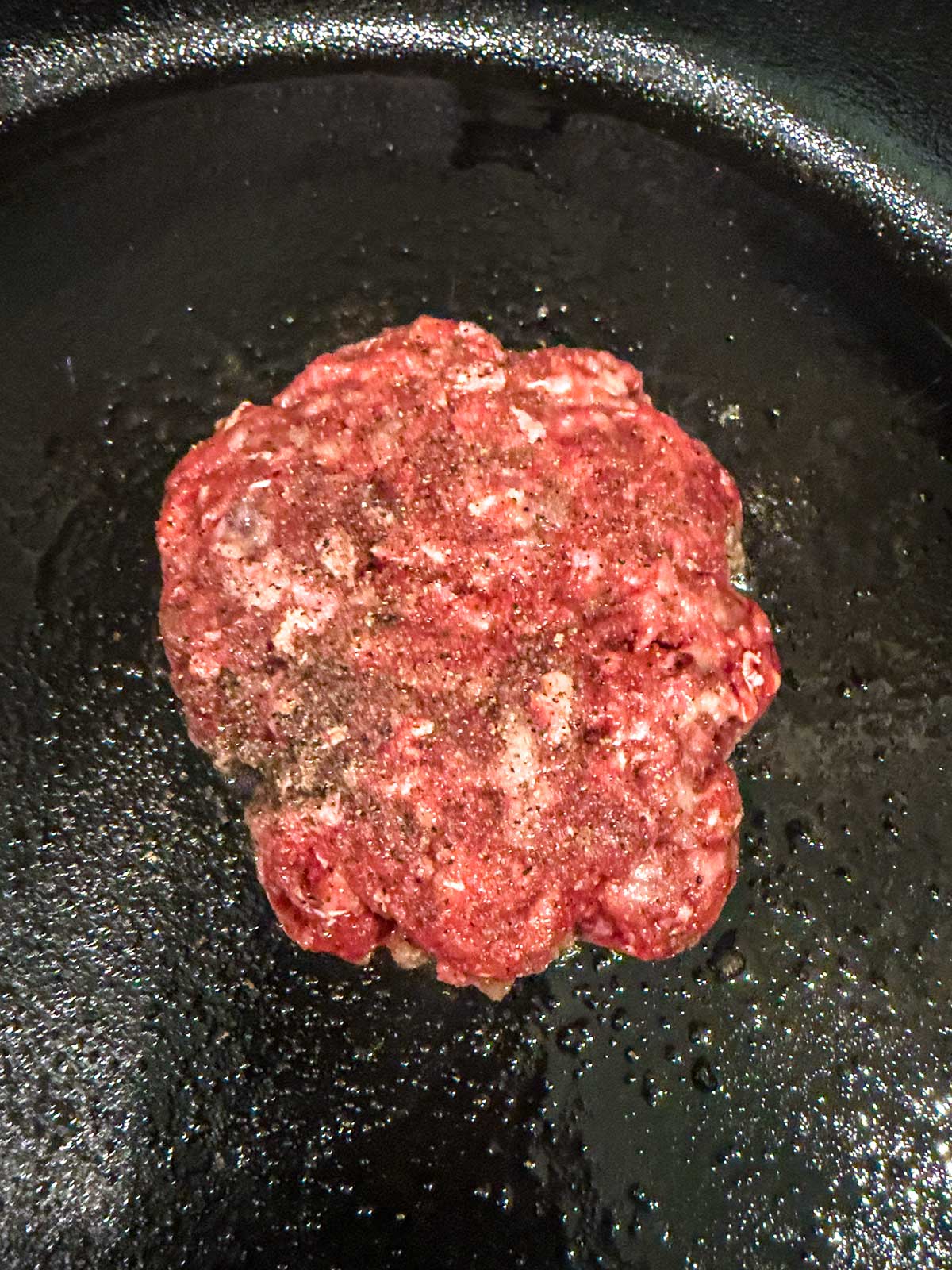 A raw, seasoned hamburger patty is cooking in a black cast iron pan, with tiny drops of oil visible around it—a classic start to delicious cast iron burgers.