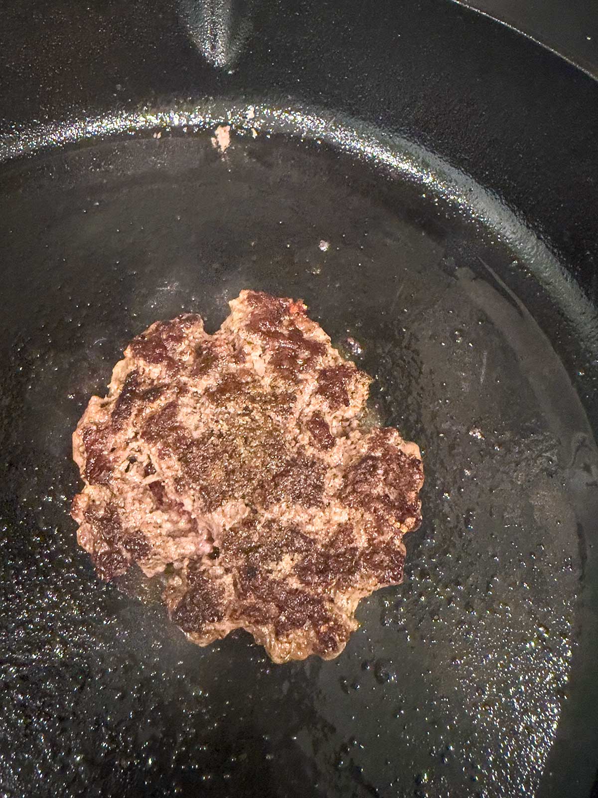 A single hamburger patty is cooking in a black cast iron skillet. The patty is browned and seared, surrounded by a small amount of oil—classic cast iron burgers in the making.