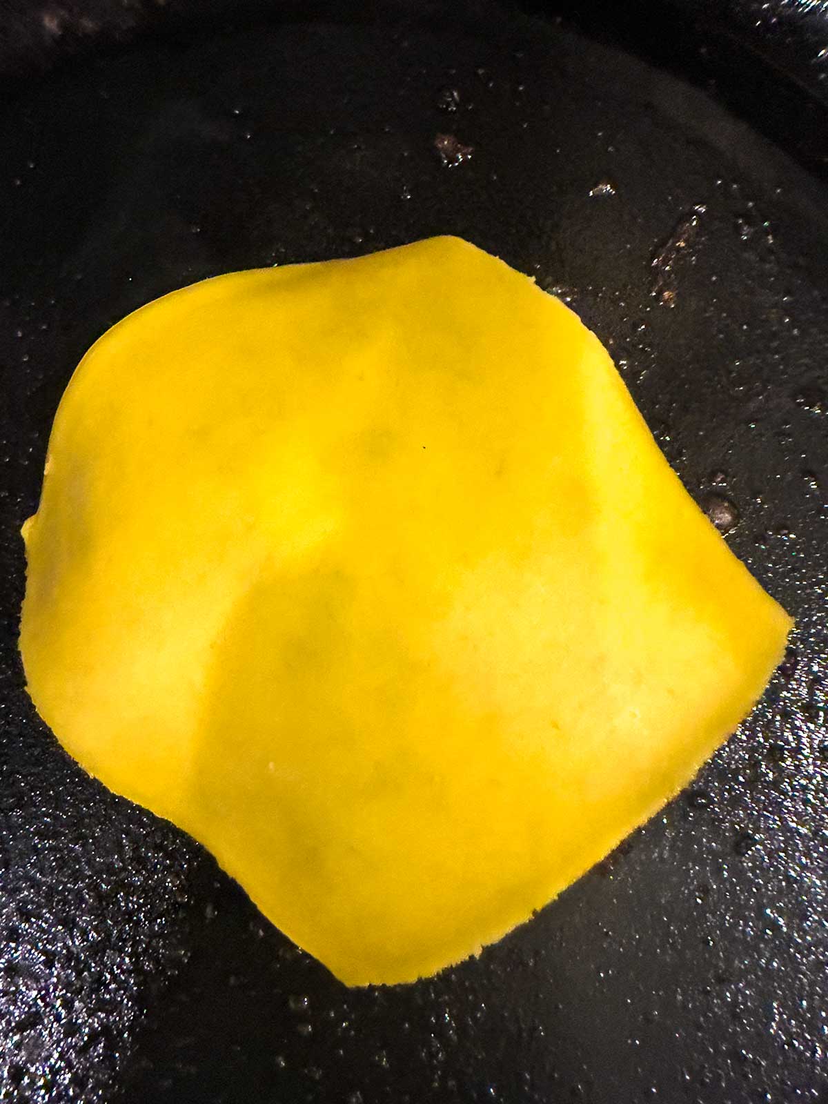 A slice of yellow American cheese melting on a black skillet, perfect for topping juicy cast iron burgers.