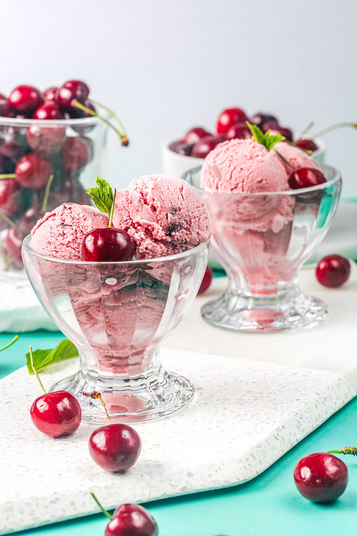 Glass bowls filled with scoops of pink cherry ice cream, garnished with fresh cherries and mint leaves. Whole cherries are scattered on the surface around the bowls.