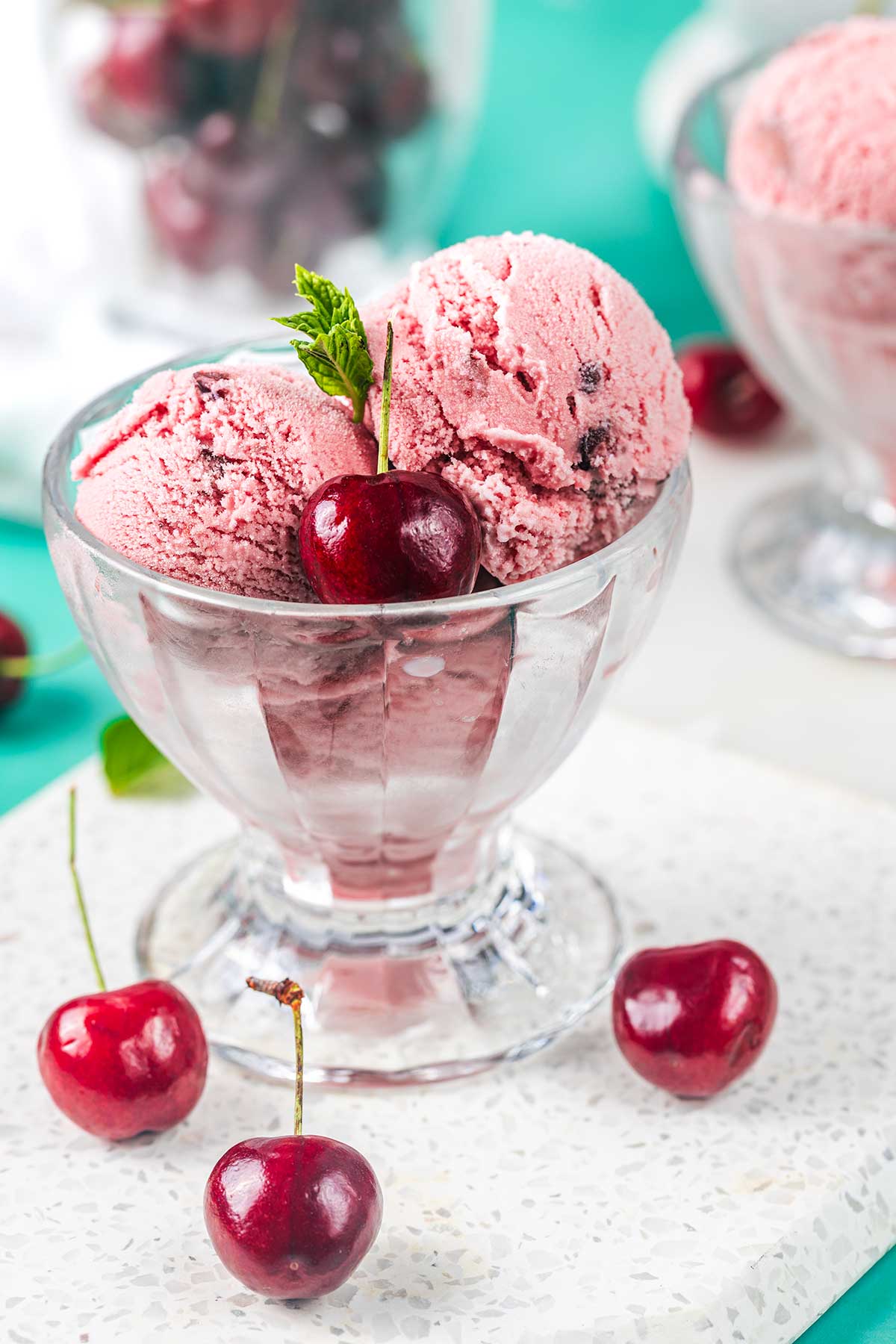 A glass bowl filled with two scoops of cherry ice cream, garnished with a fresh cherry and mint leaves. Whole cherries are scattered around the bowl on a white surface.