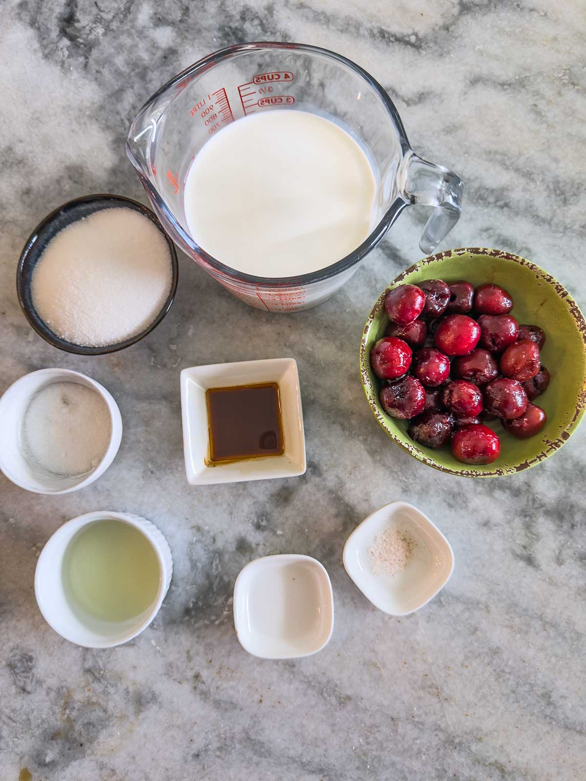 A top-down view of cherry ice cream ingredients on a marble surface, including a measuring cup of milk, a bowl of frozen cherries, sugar, salt, vanilla extract, lemon juice, and two small white dishes with powders.