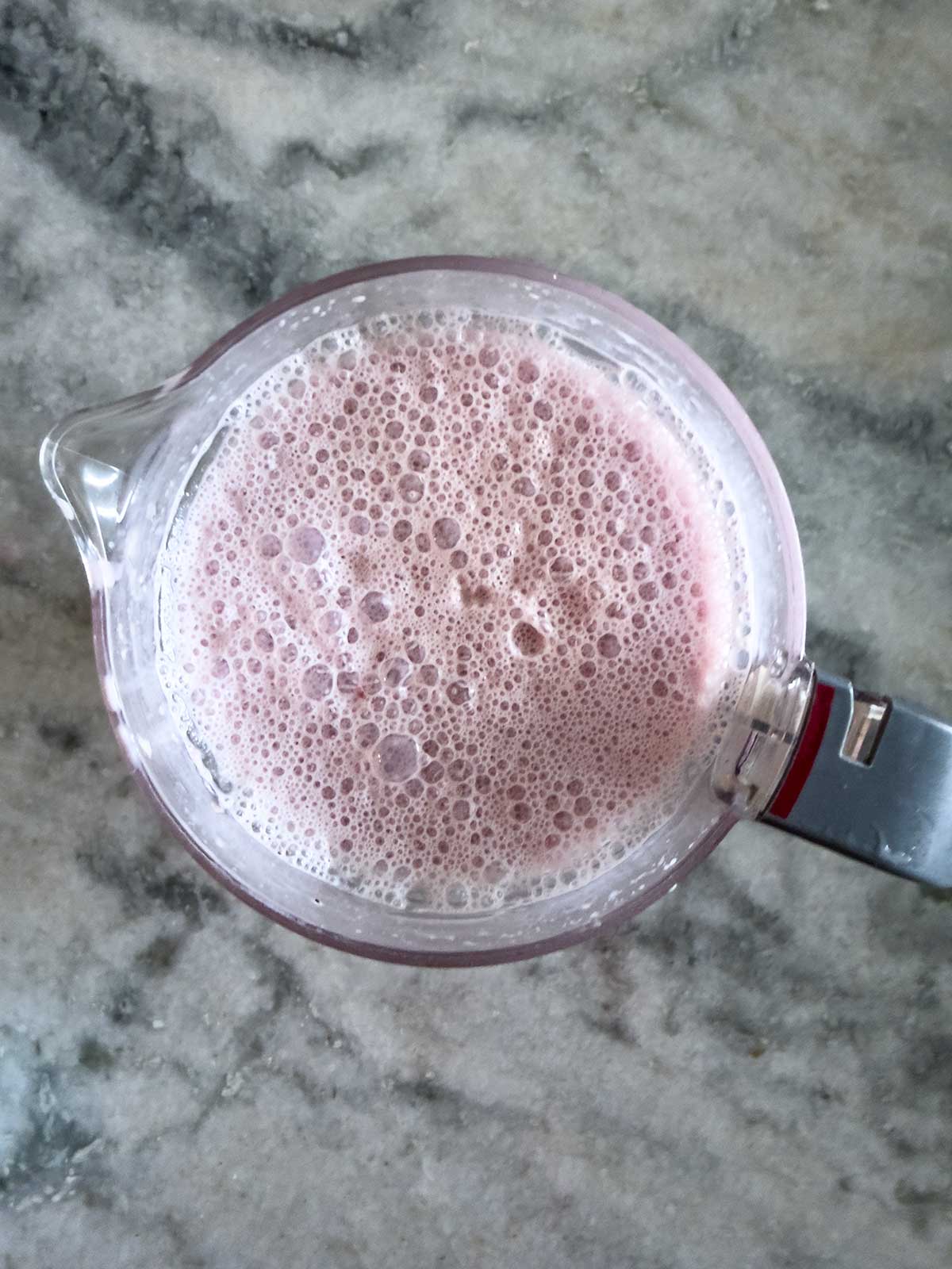 A blender filled with a frothy pink liquid, reminiscent of melted cherry ice cream, sits on a gray marble countertop, viewed from above.