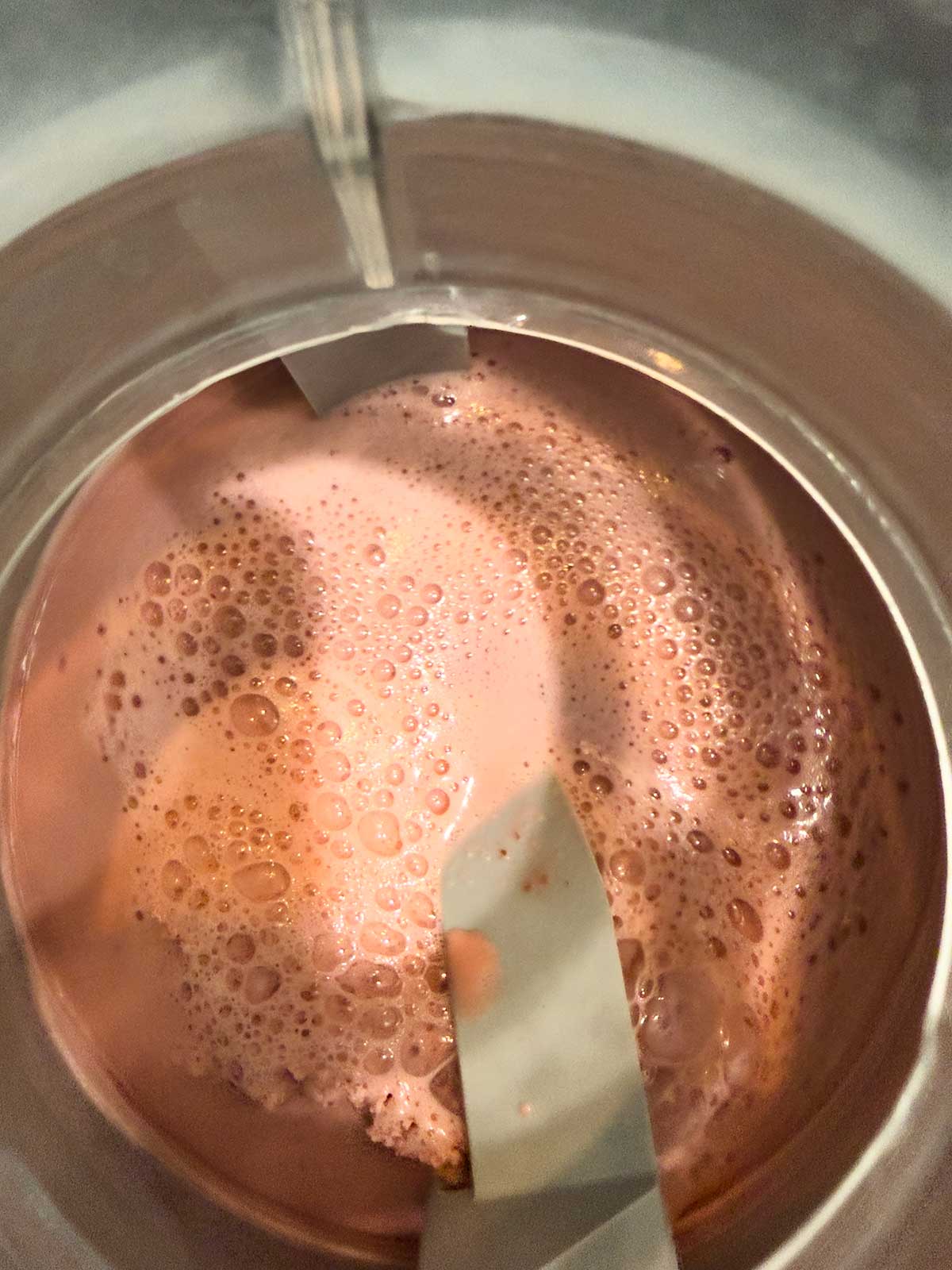Close-up of cherry ice cream mixture churning inside an ice cream maker, with bubbles and smooth texture visible on the surface. Two plastic paddles are partially submerged in the creamy mixture.