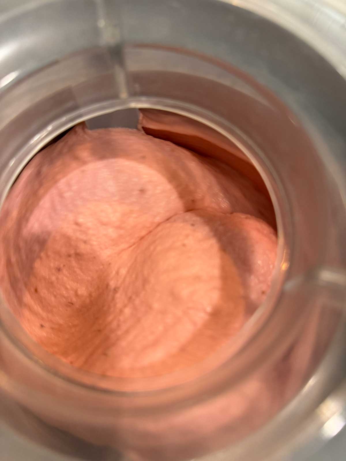 Close-up view from above of a pink, foamy cherry ice cream mixture inside a clear container, possibly a blender or food processor. The texture appears smooth and airy.