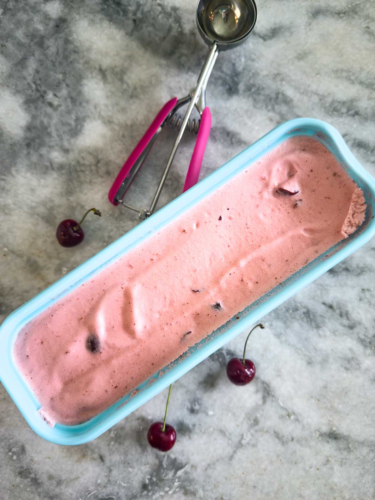 A rectangular blue container holds cherry ice cream with visible cherry pieces. A metal ice cream scoop with a pink handle and three fresh cherries rest on the gray marble surface nearby.