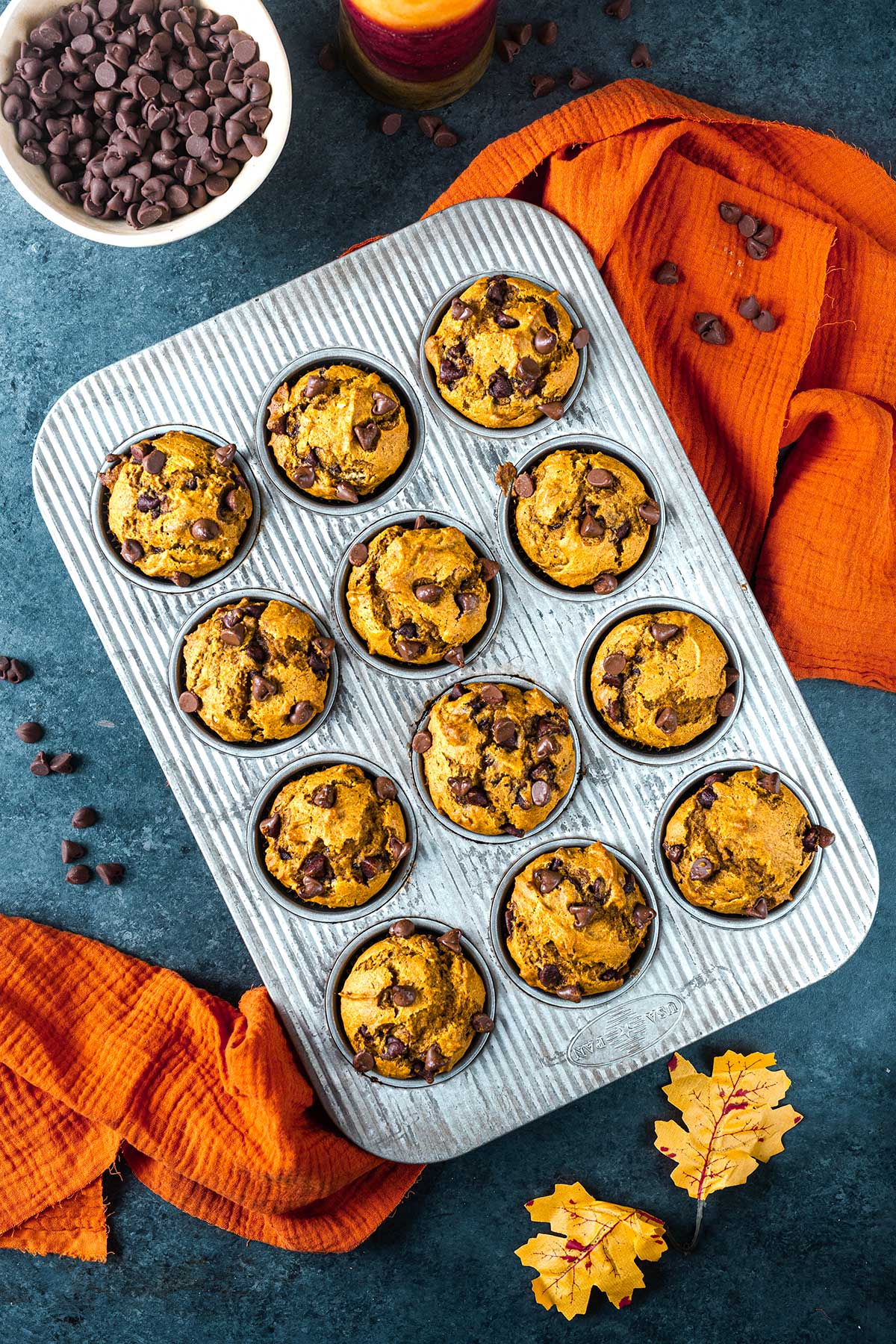 A muffin tin filled with pumpkin chocolate chip muffins sits on a dark blue surface with scattered chocolate chips, orange cloths, and autumn leaves; a bowl of chocolate chips, a candle, and Ninja Creami Pumpkin Ice Cream are nearby.