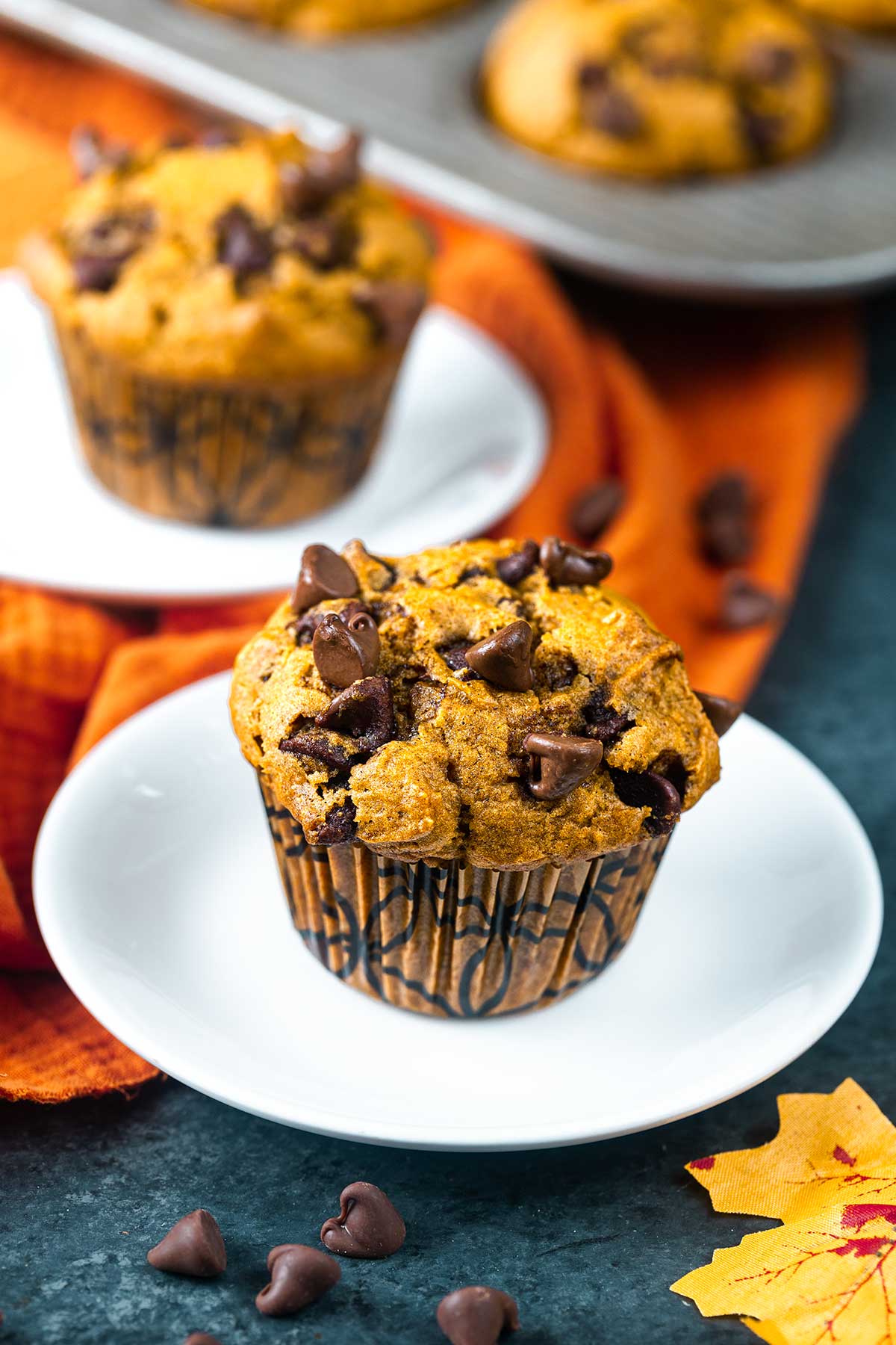 A pumpkin chocolate chip muffin sits on a white plate, topped with chocolate chips. In the background, Ninja Creami Pumpkin Ice Cream is paired with another muffin, while orange fabric and scattered chocolate chips complete the cozy scene.