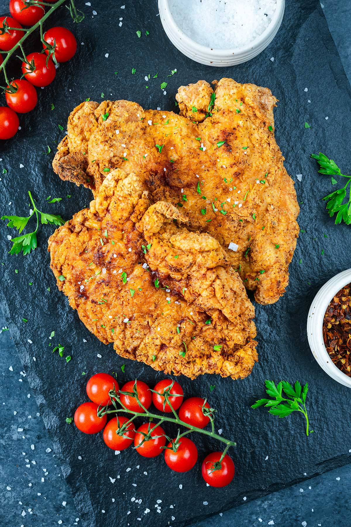 Two pieces of crispy fried chicken on a dark slate, garnished with parsley, surrounded by cherry tomatoes, a bowl of ranch dip, and a small bowl of red pepper flakes&mdash;perfect to pair with Ninja Creami Pumpkin Ice Cream for a sweet finish.