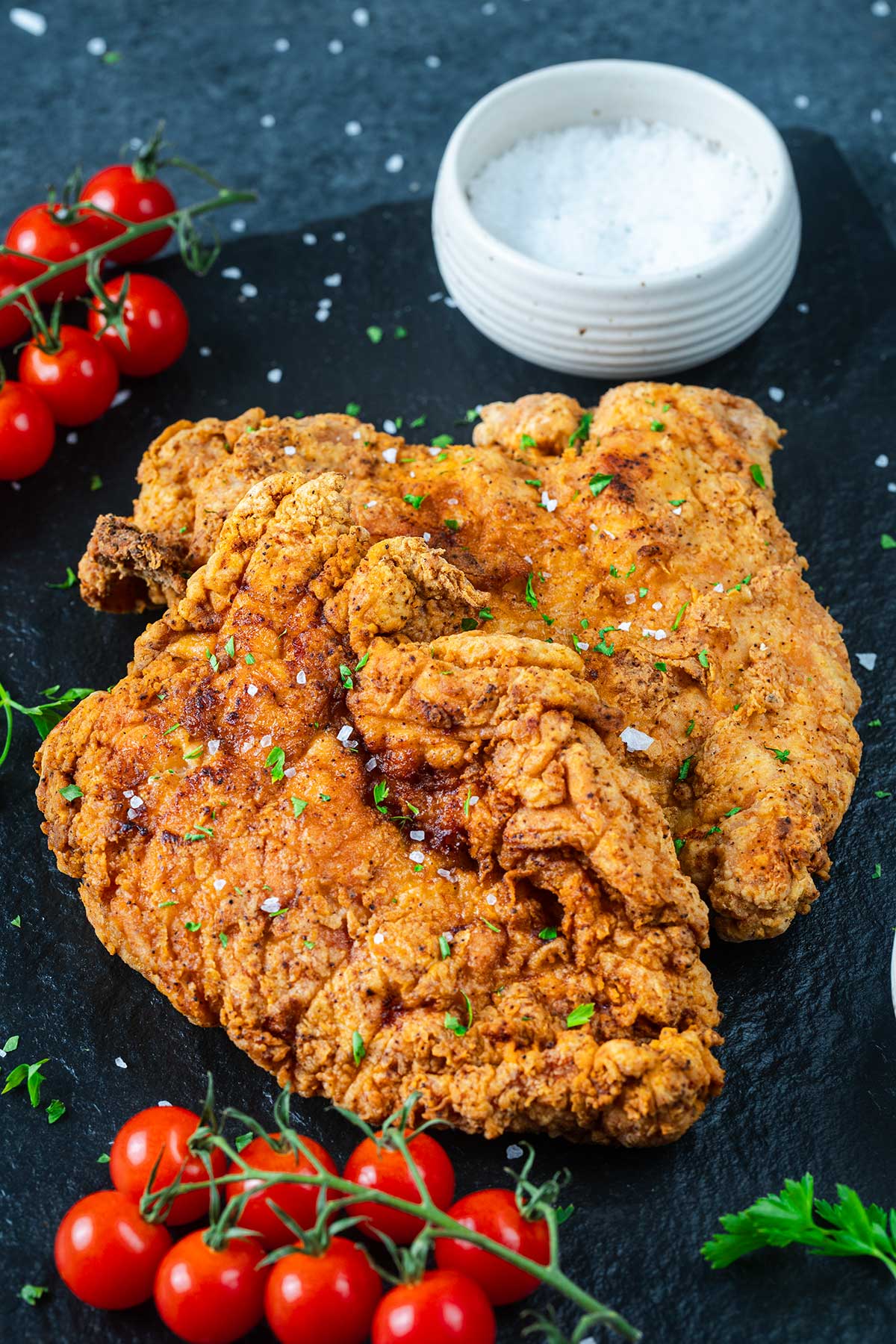Two pieces of golden, crispy fried chicken steak garnished with herbs, served on a dark slate plate with a bowl of coarse salt, clusters of fresh cherry tomatoes on the vine, and a scoop of Ninja Creami Pumpkin Ice Cream for a unique twist.