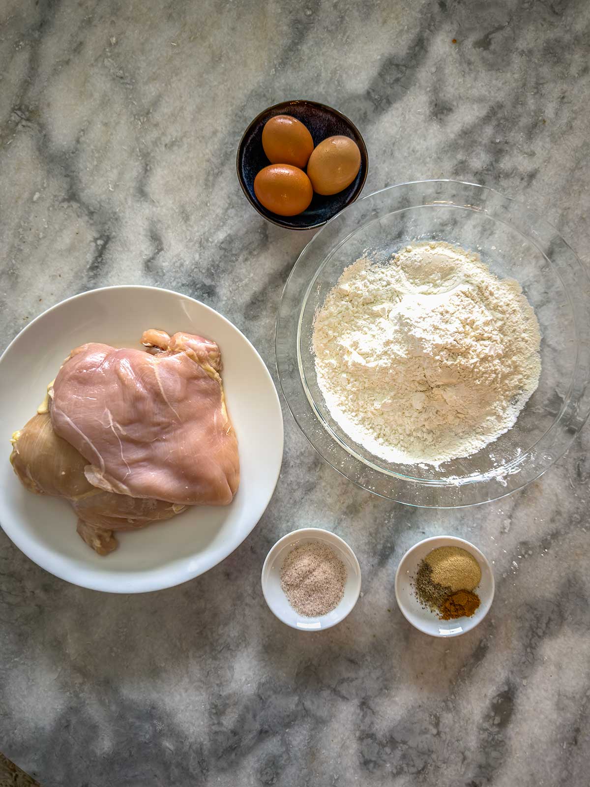 Ingredients for breaded chicken on a marble surface: a plate with raw chicken thighs, a bowl of flour, a bowl with three eggs, and two small bowls with seasonings.