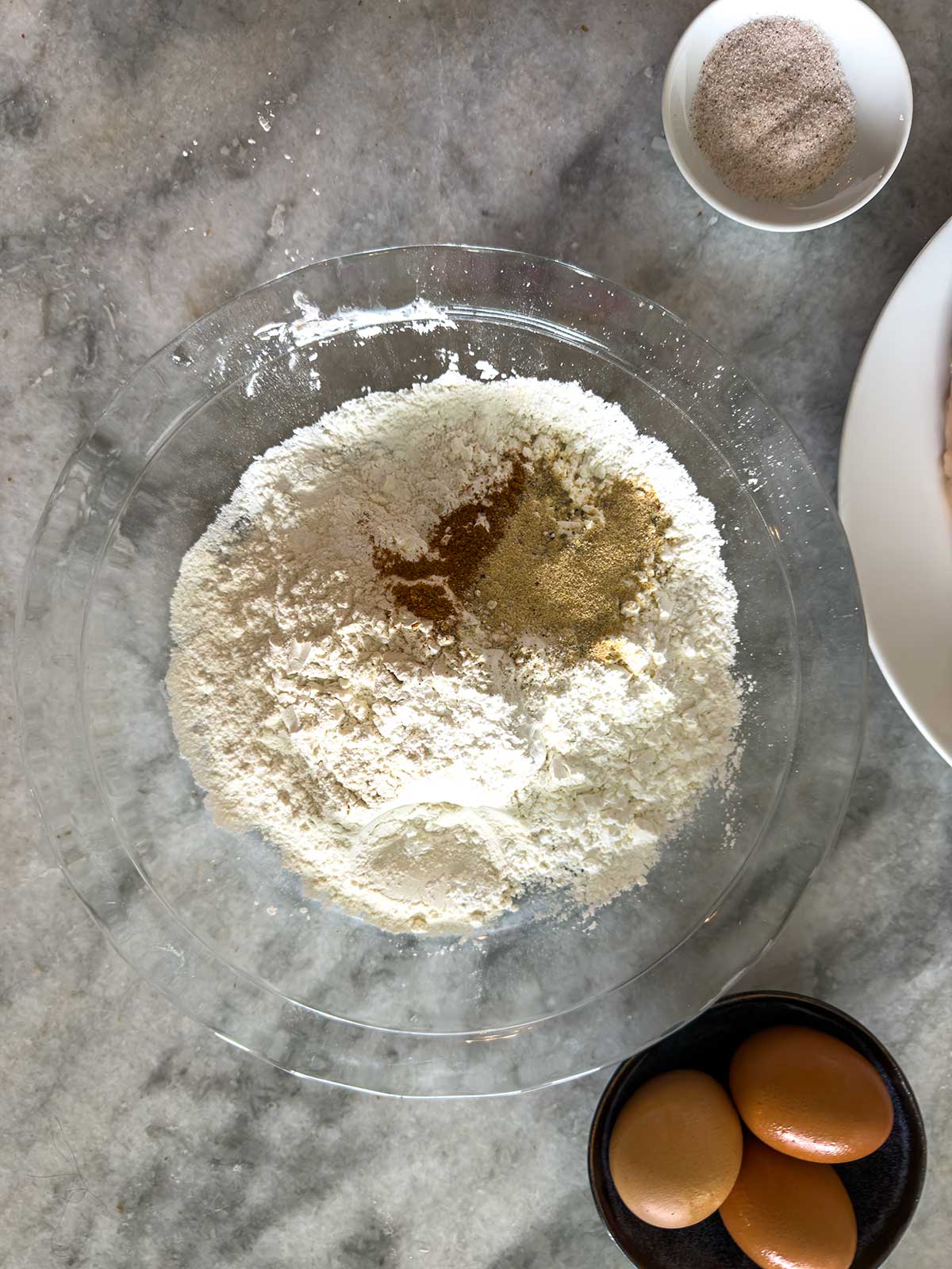 A glass mixing bowl with flour, black pepper, and other spices sits on a marble countertop, surrounded by a small bowl of eggs and a small dish of seasoned salt.