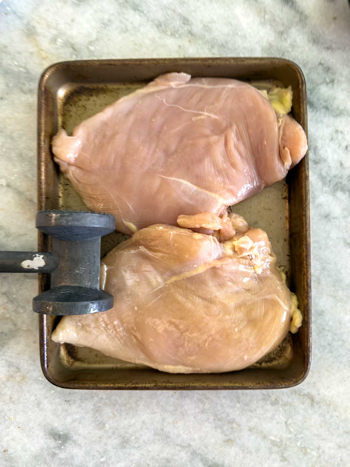 Two raw chicken breasts on a metal tray, with a meat mallet resting on the edge, placed on a light-colored countertop.