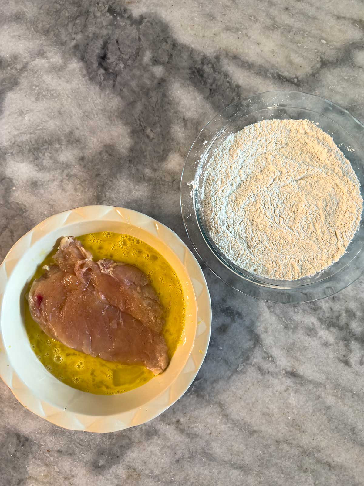 A raw chicken breast sits in a bowl with beaten eggs next to a glass bowl filled with flour on a gray marble countertop, prepared for breading.