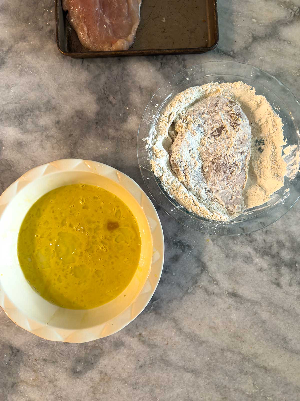 A bowl of beaten eggs and a bowl of flour with a piece of raw chicken coated in flour are on a marble countertop. An additional raw chicken piece is on a baking tray nearby.
