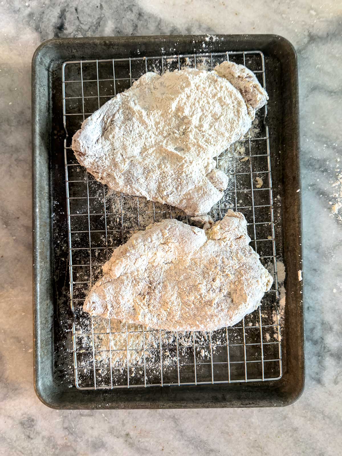Two pieces of raw, flour-coated chicken breasts rest on a wire rack set over a baking tray. The tray is placed on a marble countertop, and some flour is scattered around.