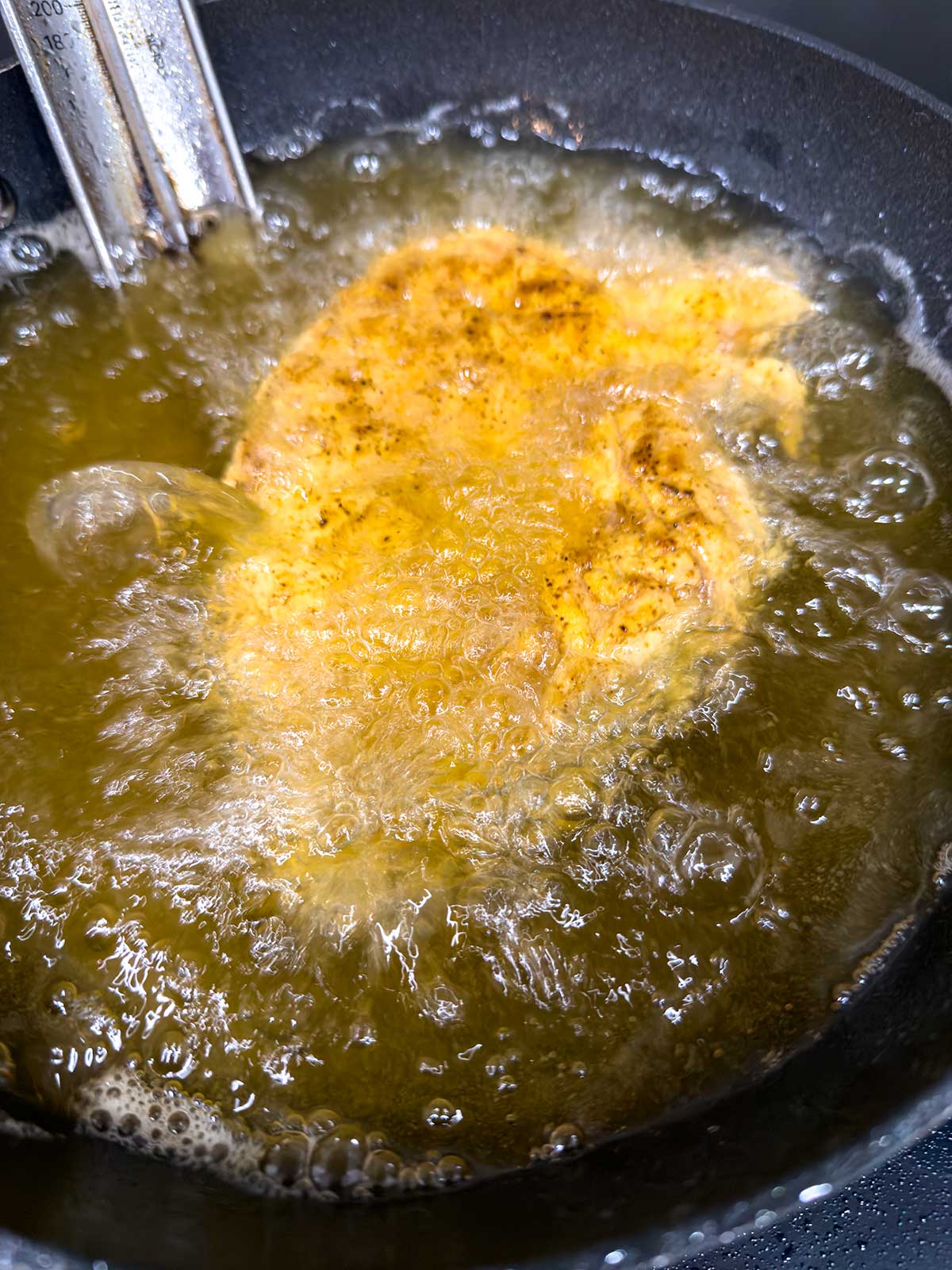 A piece of seasoned chicken is being deep-fried in hot oil inside a black frying pan, with bubbles surrounding the chicken as it cooks.