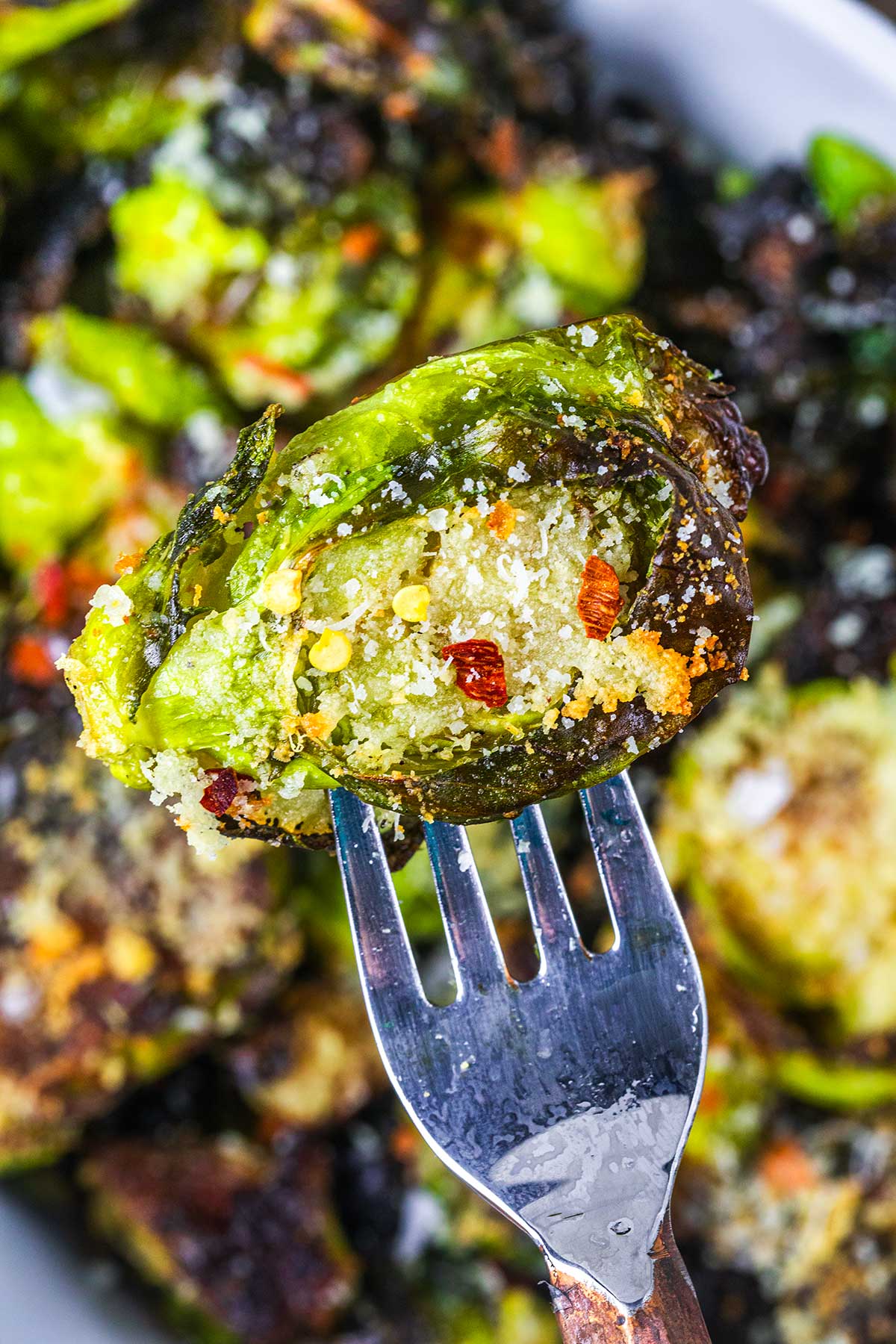 A close-up of a fork holding a crispy, smashed Brussels sprout topped with grated parmesan cheese, red pepper flakes, and coarse salt, with more Brussels sprouts in the blurred background.