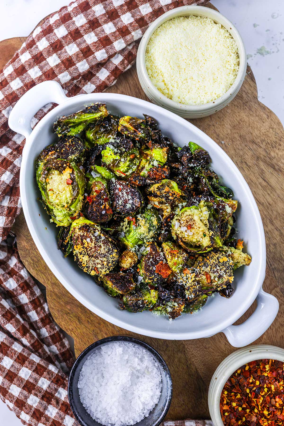 Roasted Brussels sprouts in a white oval dish, topped with grated cheese and red pepper flakes, with sides of grated cheese, coarse salt, and red pepper flakes on a wooden board with a brown checkered cloth.