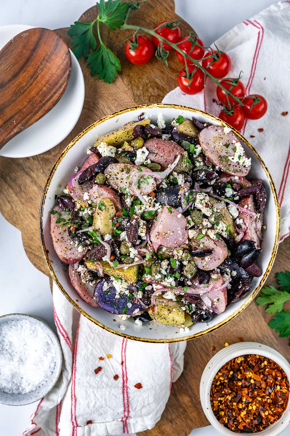 A bowl of potato salad with black beans, capers, feta cheese, and herbs sits on a wooden board, alongside fresh parsley, cherry tomatoes, sautéed mushrooms for steak, a bowl of salt, and red pepper flakes.